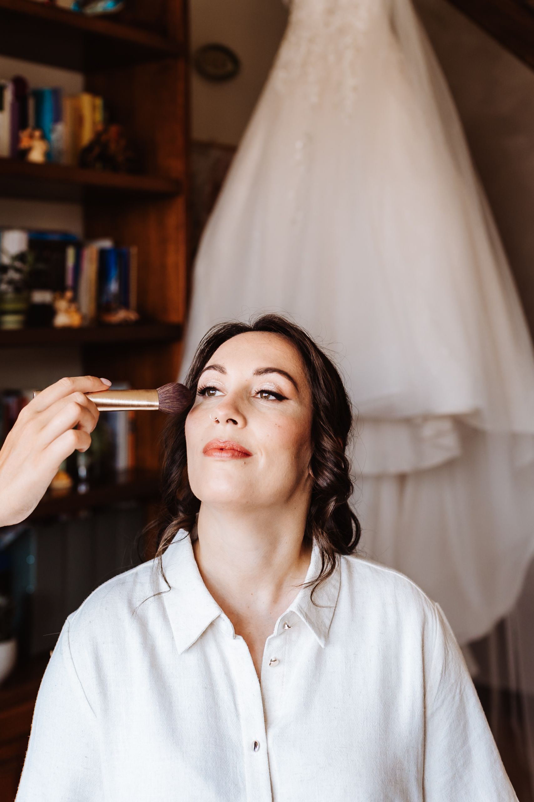 Bride getting makeup applied with wedding dress hanging in background during bridal preparation.