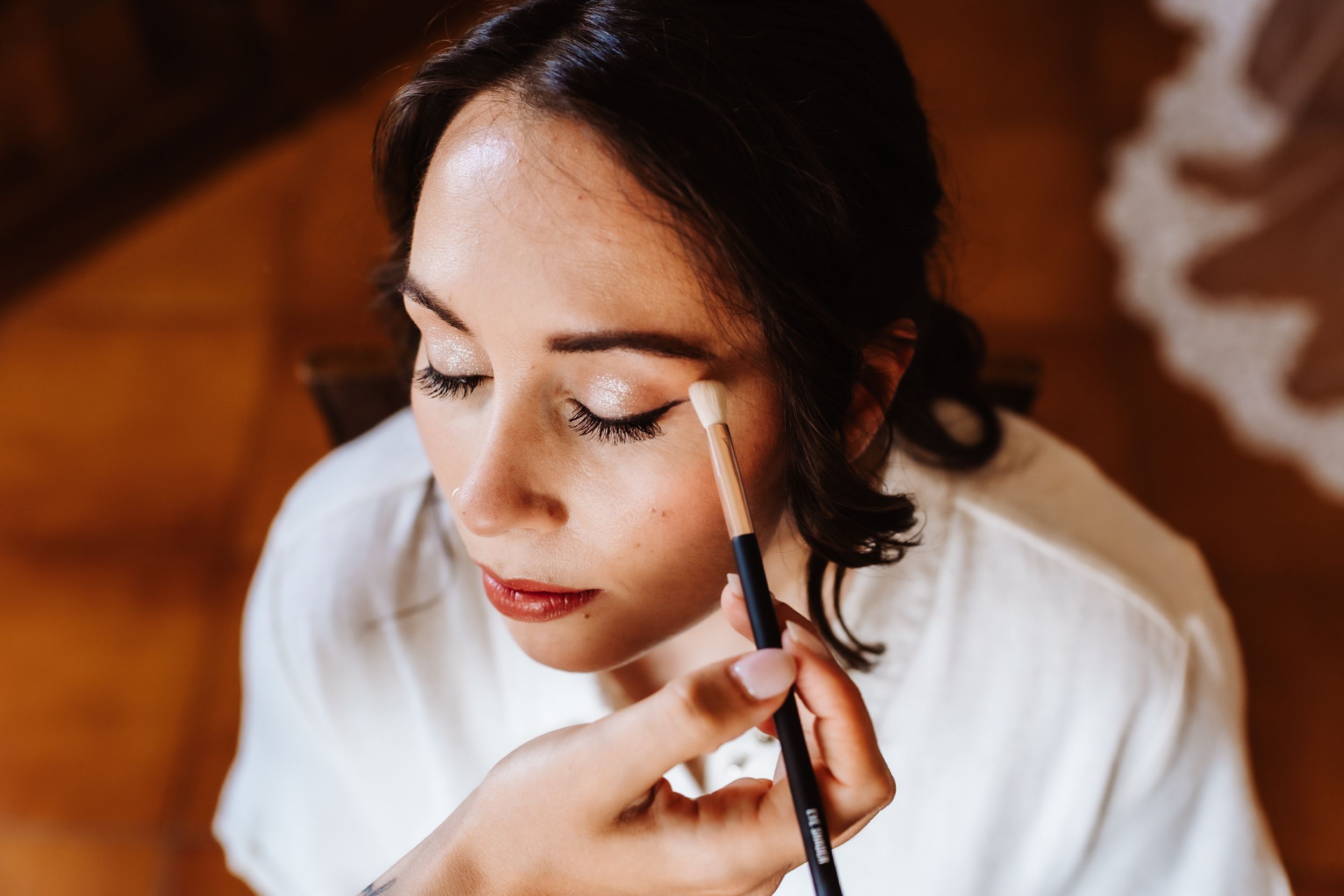 A makeup artist applies eyeshadow with a brush to a brunette woman in a white robe during bridal preparation.