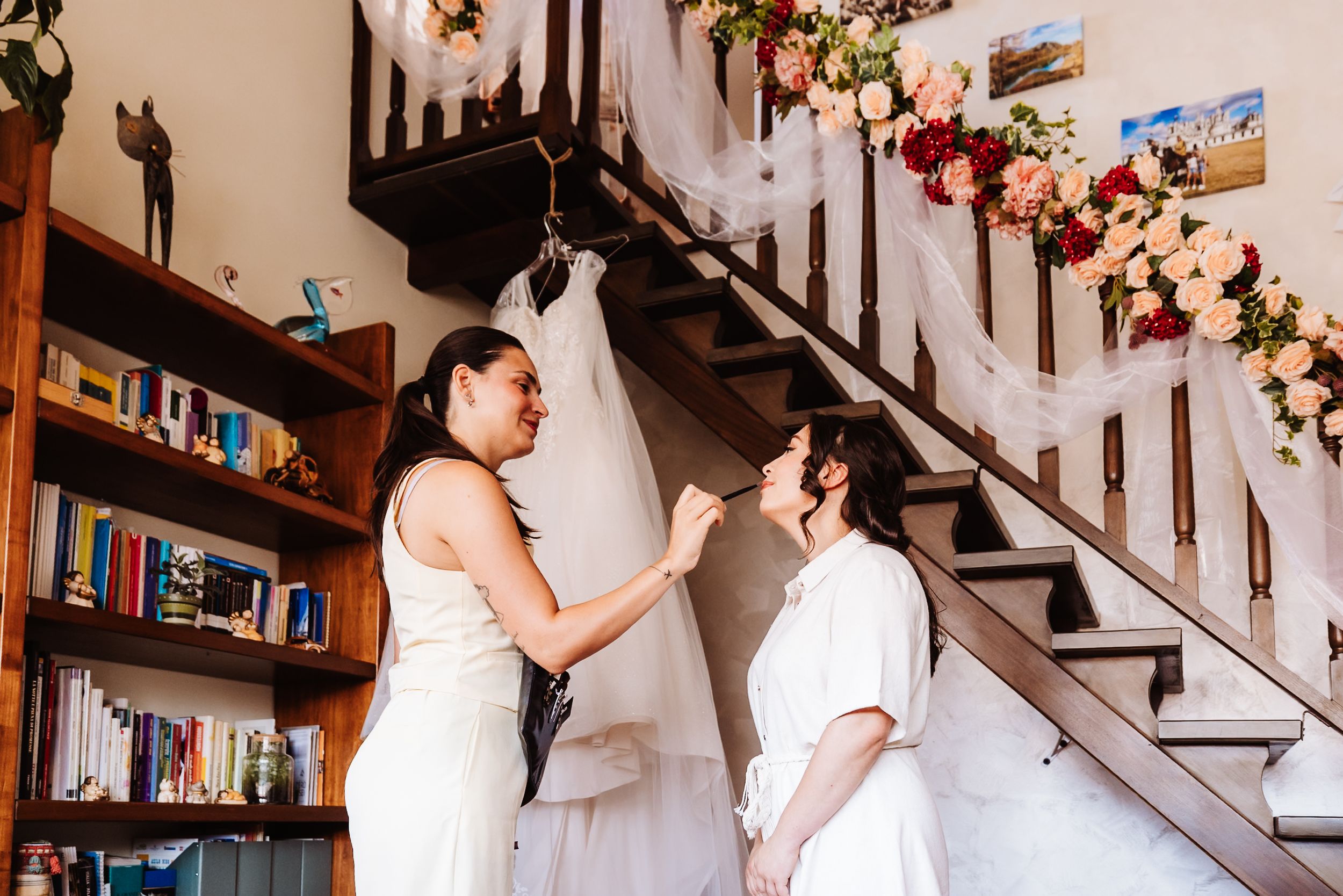 Two brides share a joyful moment by flower-adorned staircase with wedding dress hanging nearby.