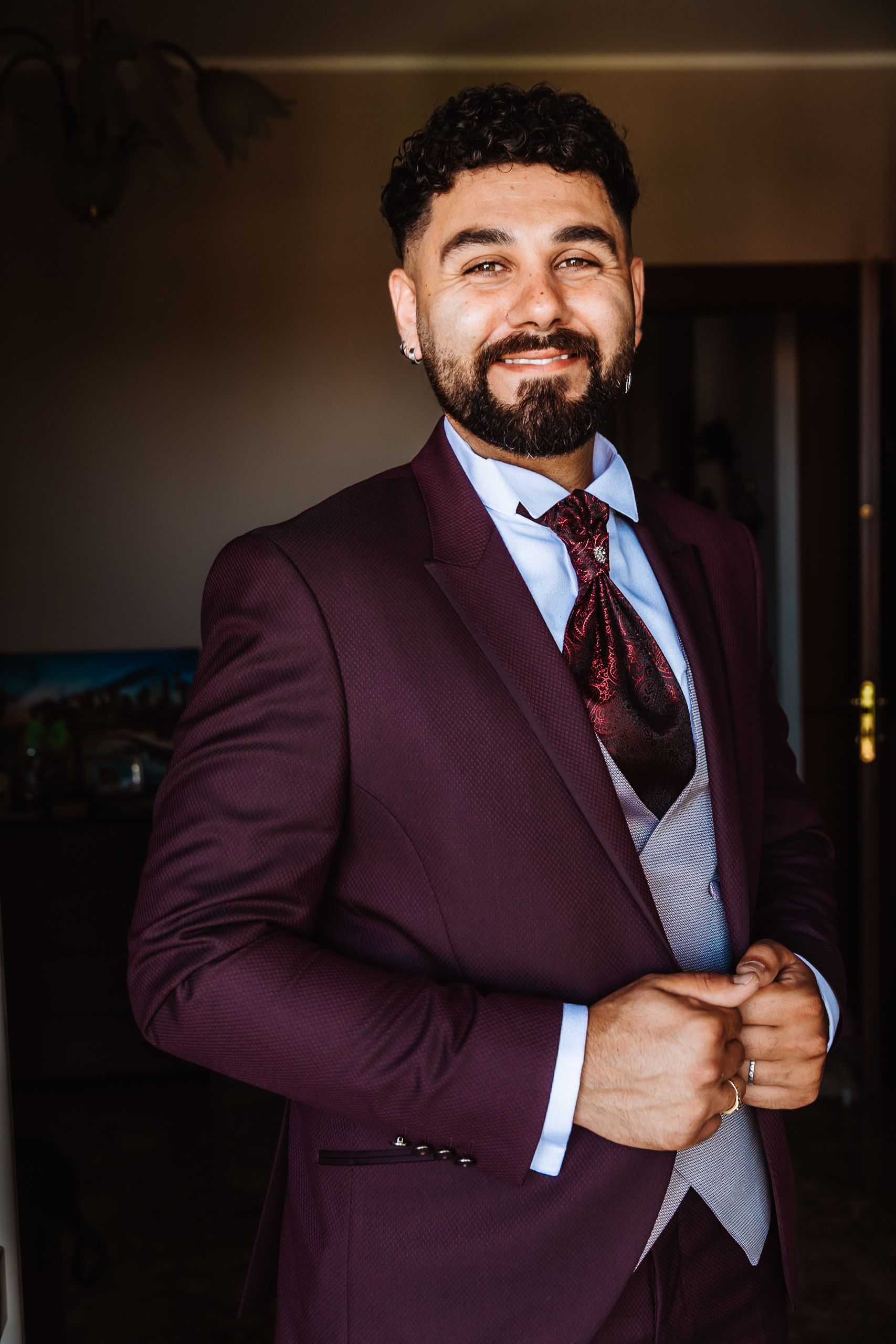 Smiling man with beard wearing a burgundy suit and floral tie, adjusting his jacket indoors.