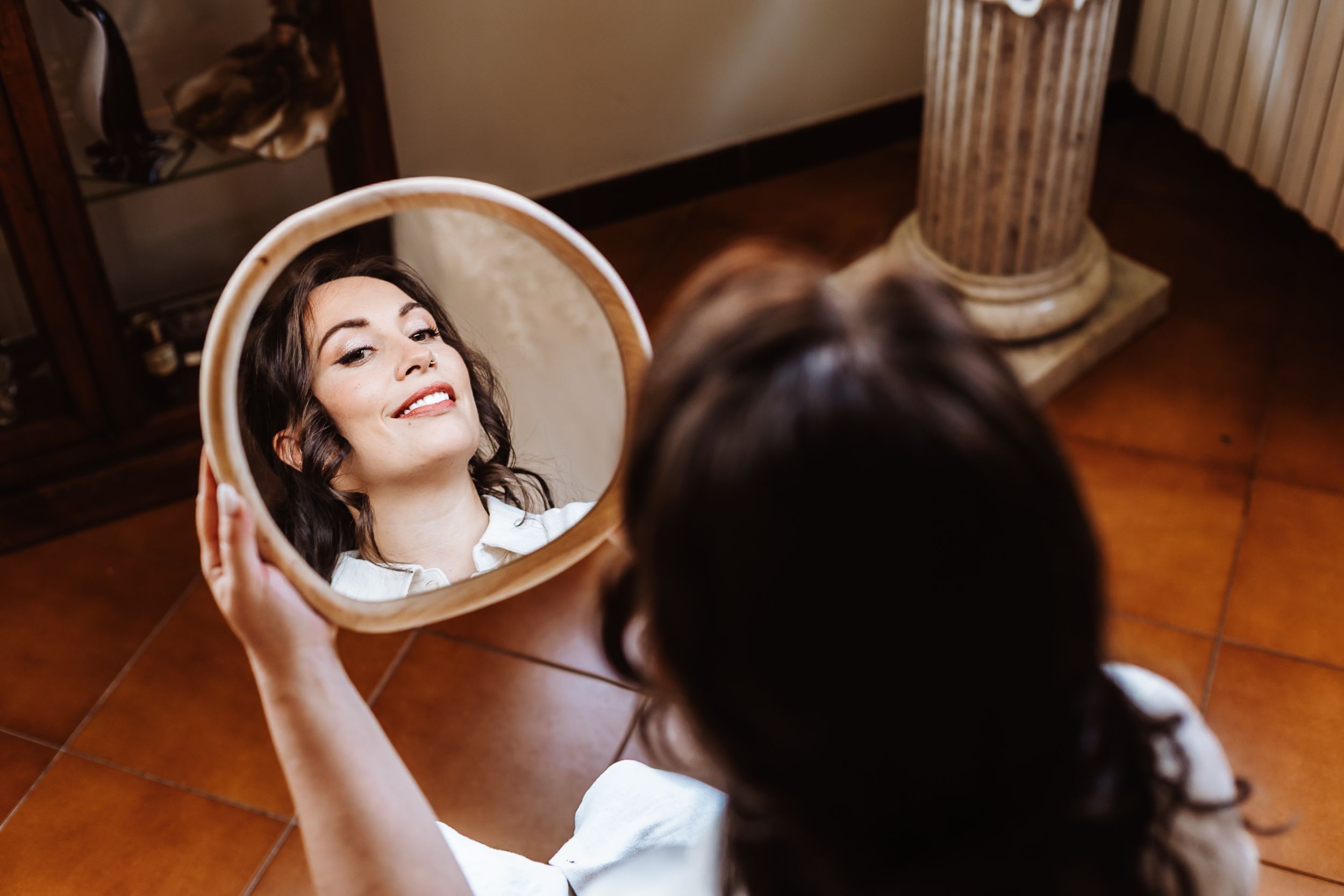 Young woman holding a round wooden mirror, smiling at her reflection in an elegant room with terracotta floors.