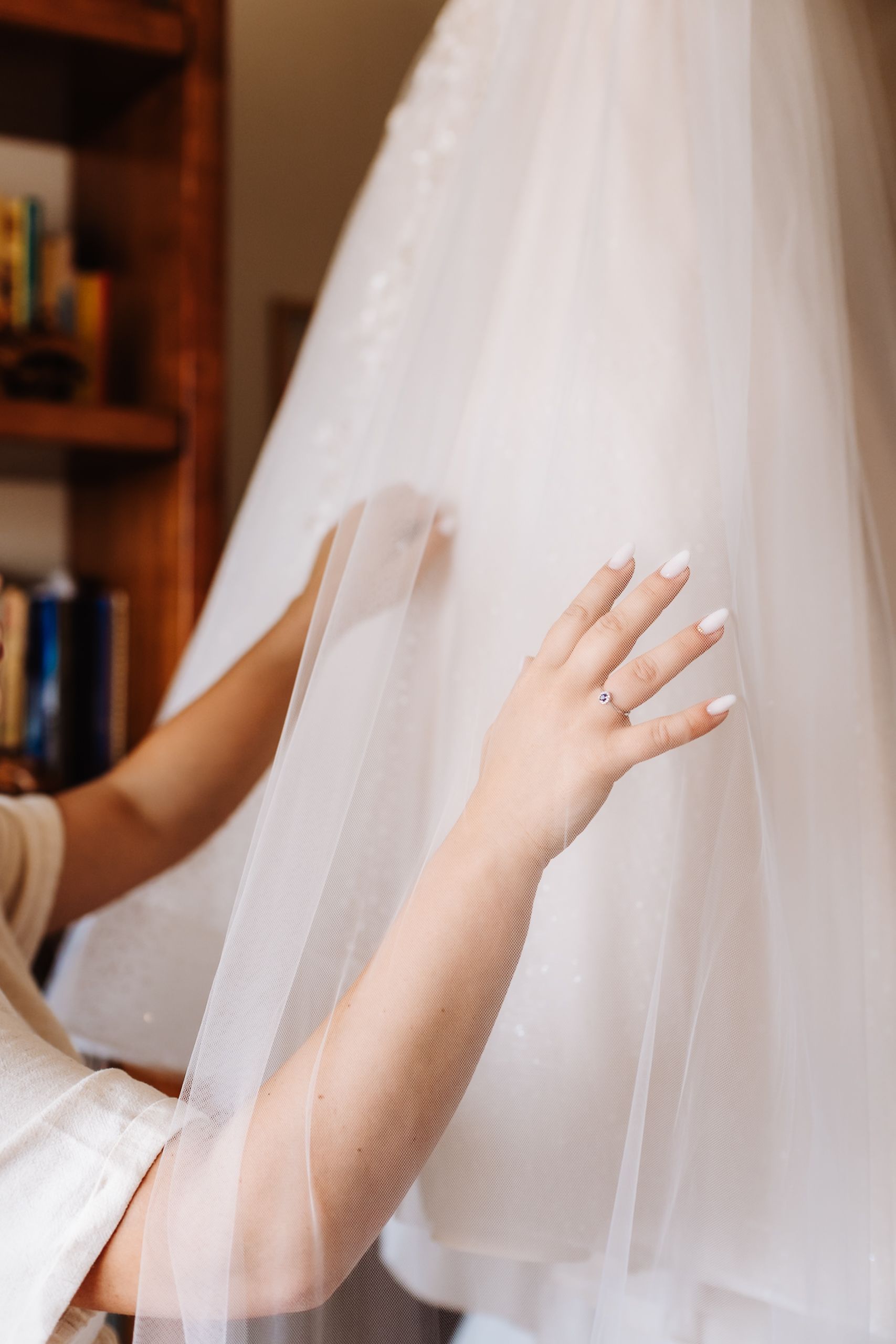 Bride touching delicate white tulle wedding veil with manicured nails and engagement ring visible.