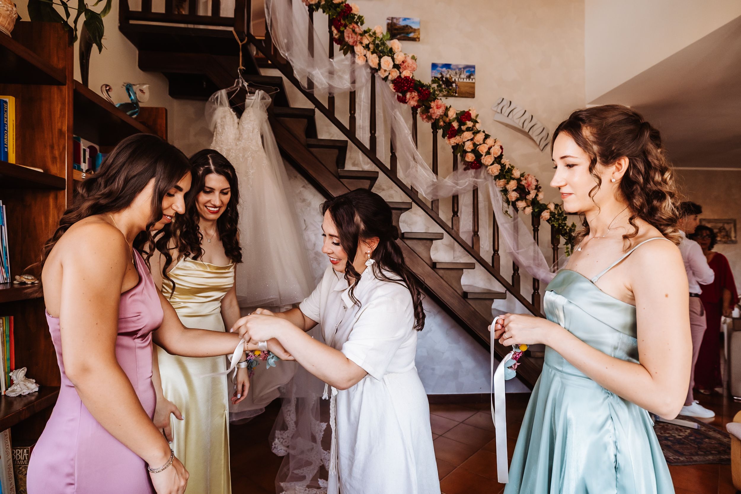 Bridesmaids in colorful dresses help bride prepare near floral-decorated staircase in rustic wedding venue.