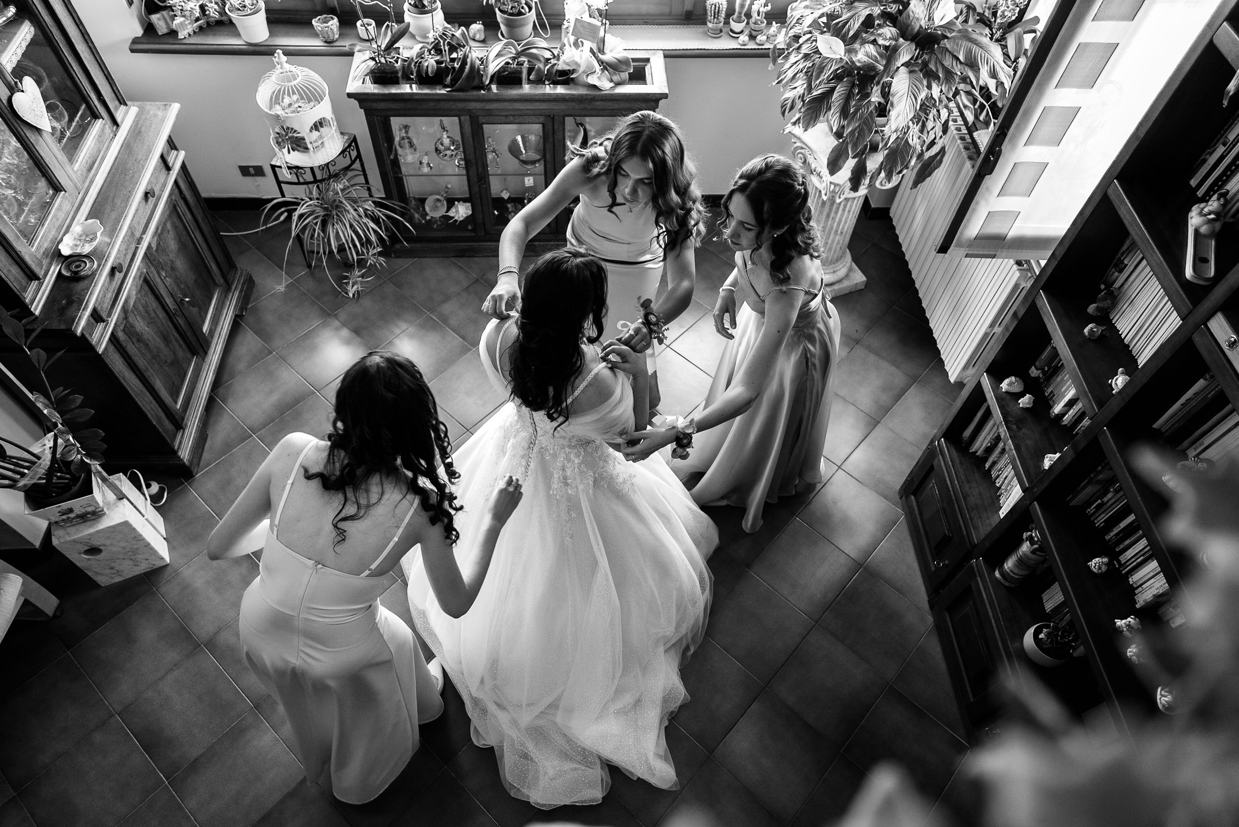 Black and white overhead shot of bride in white gown surrounded by bridesmaids helping her prepare for wedding.