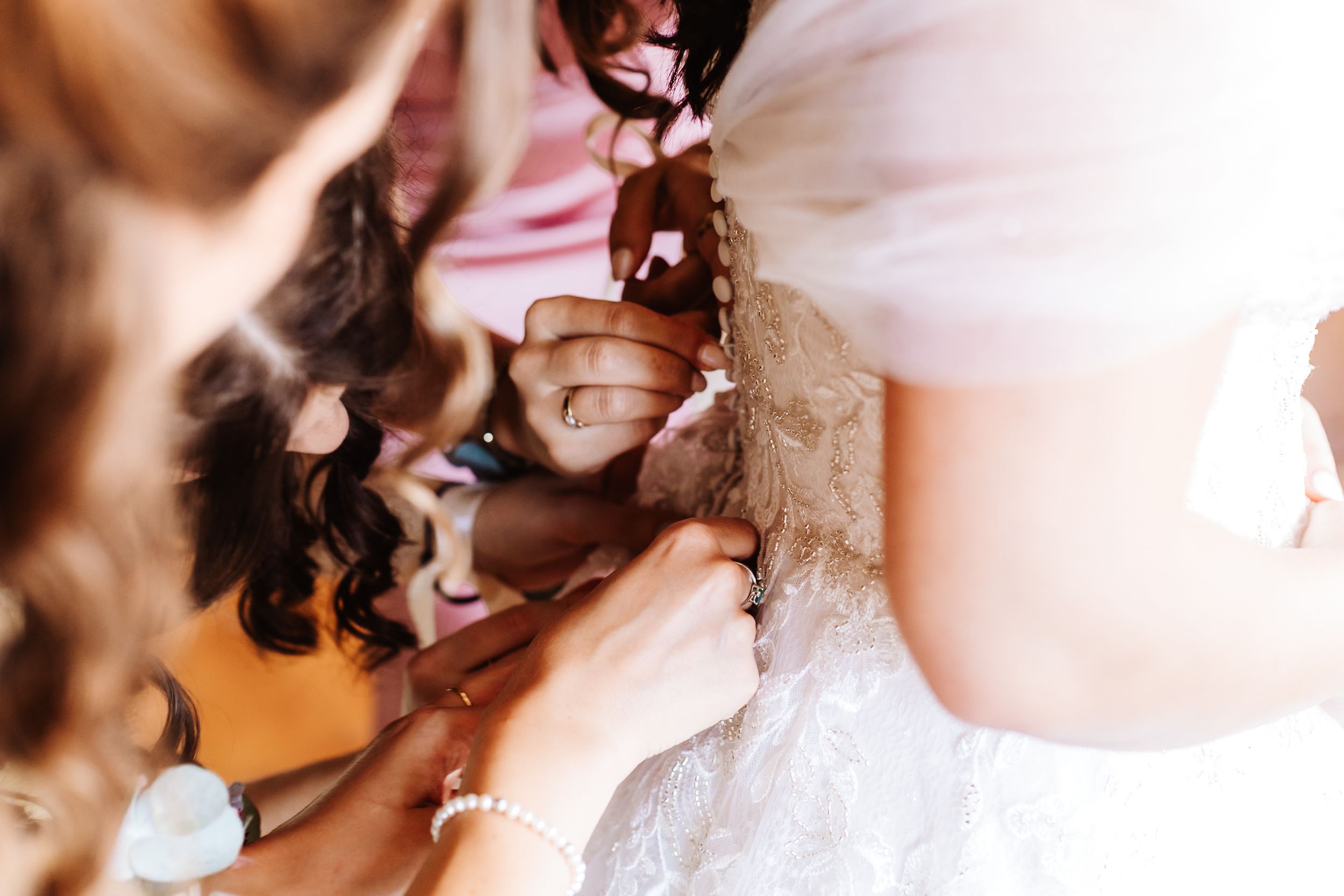 Bridesmaids helping bride button up her white lace wedding dress before the ceremony.
