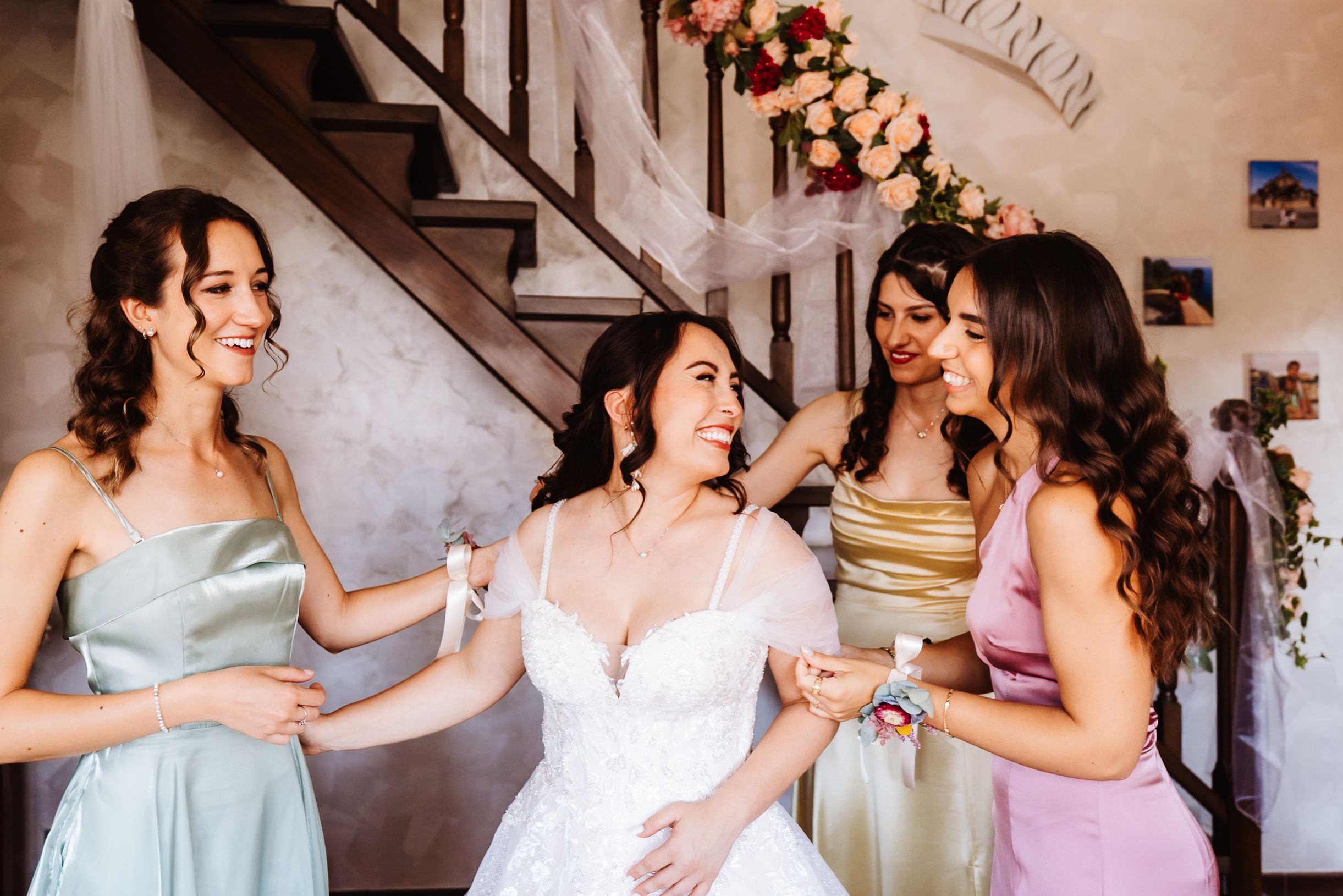 Bride in white gown laughing with three bridesmaids in colorful satin dresses near floral staircase.
