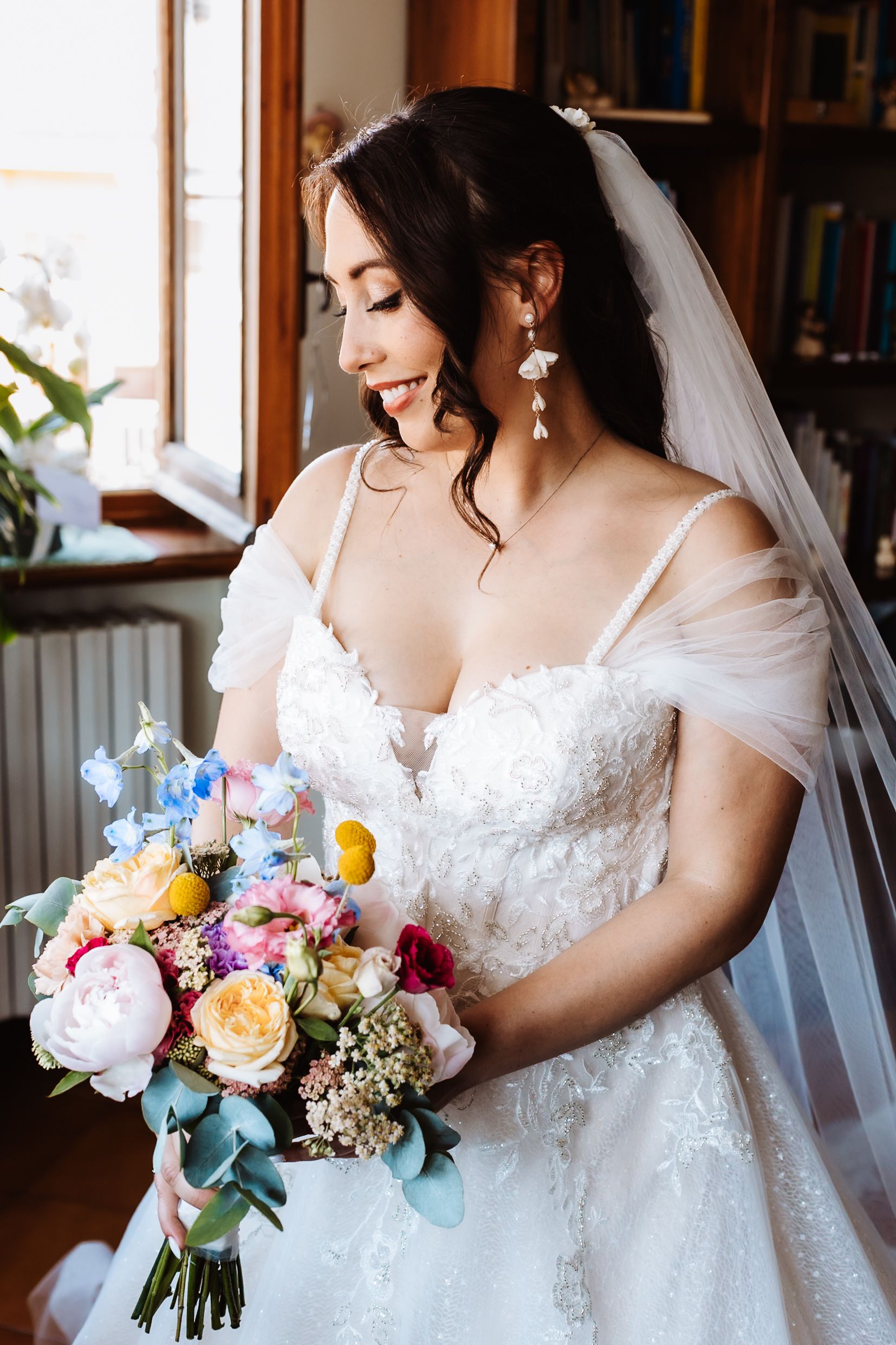 Smiling bride in lace wedding gown and veil holds colorful bouquet with peonies and roses near window.