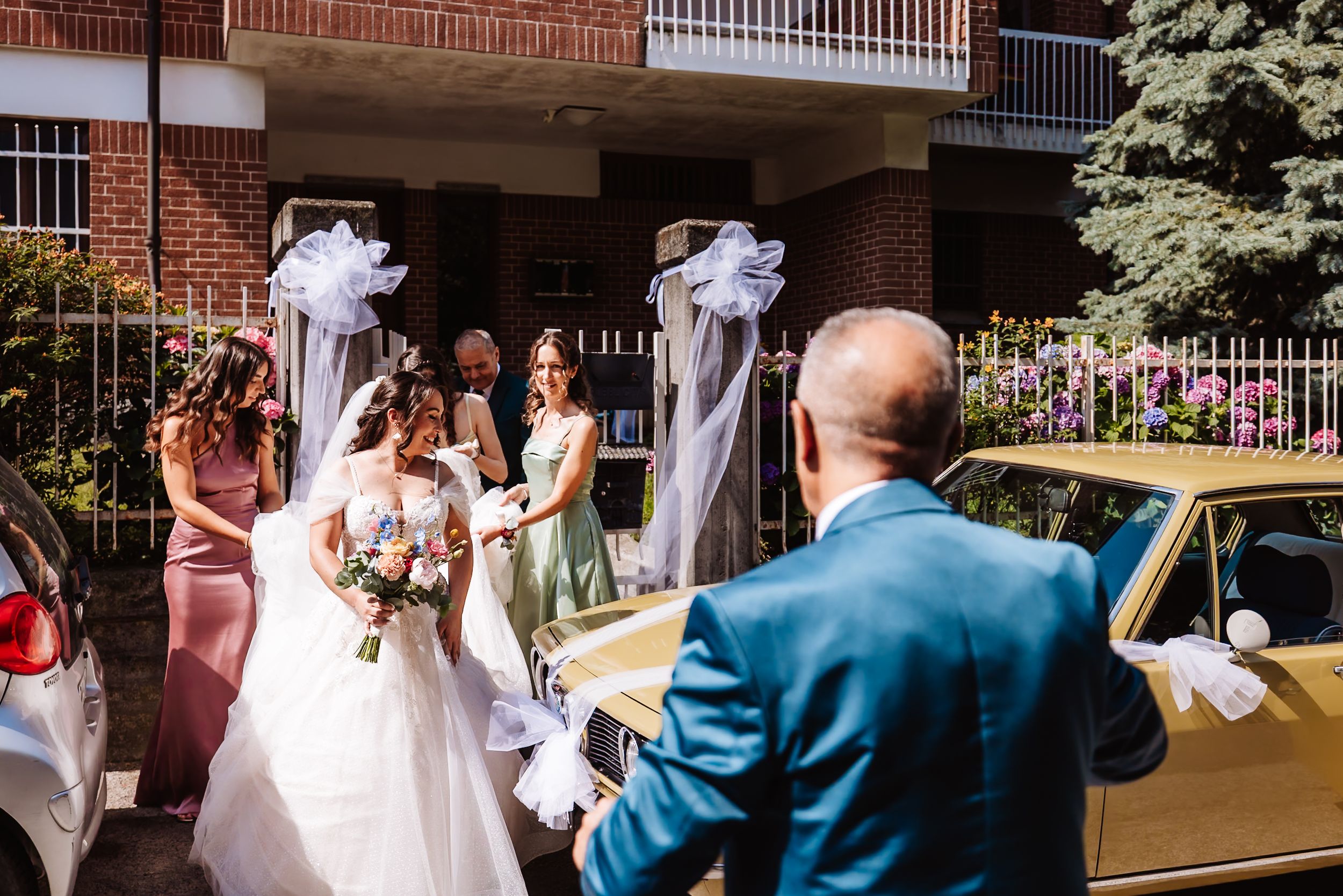 Bride in white gown holds bouquet with bridesmaids outside brick building near decorated gold wedding car.