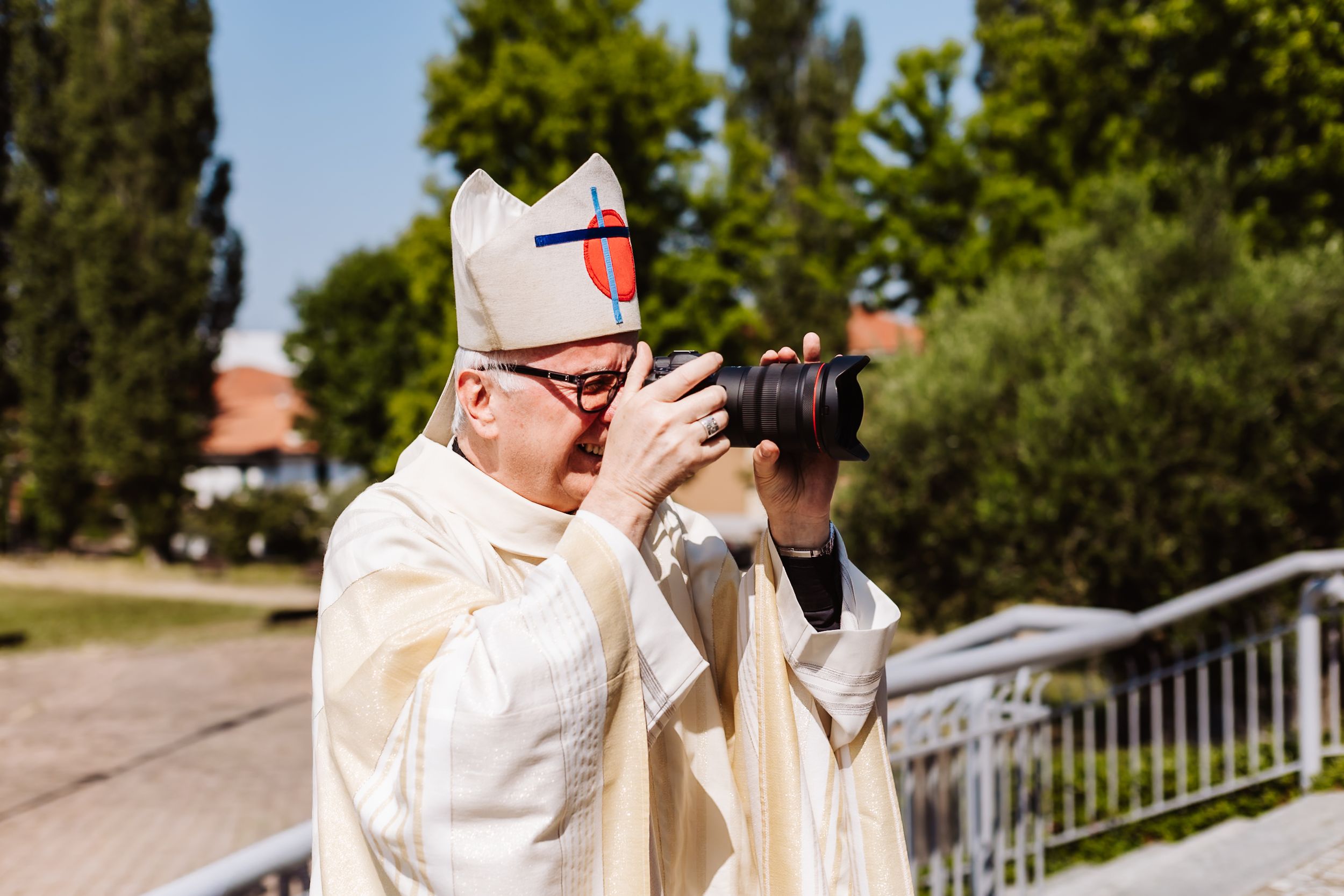A bishop in white vestments and mitre photographs with a DSLR camera outdoors on a sunny day.