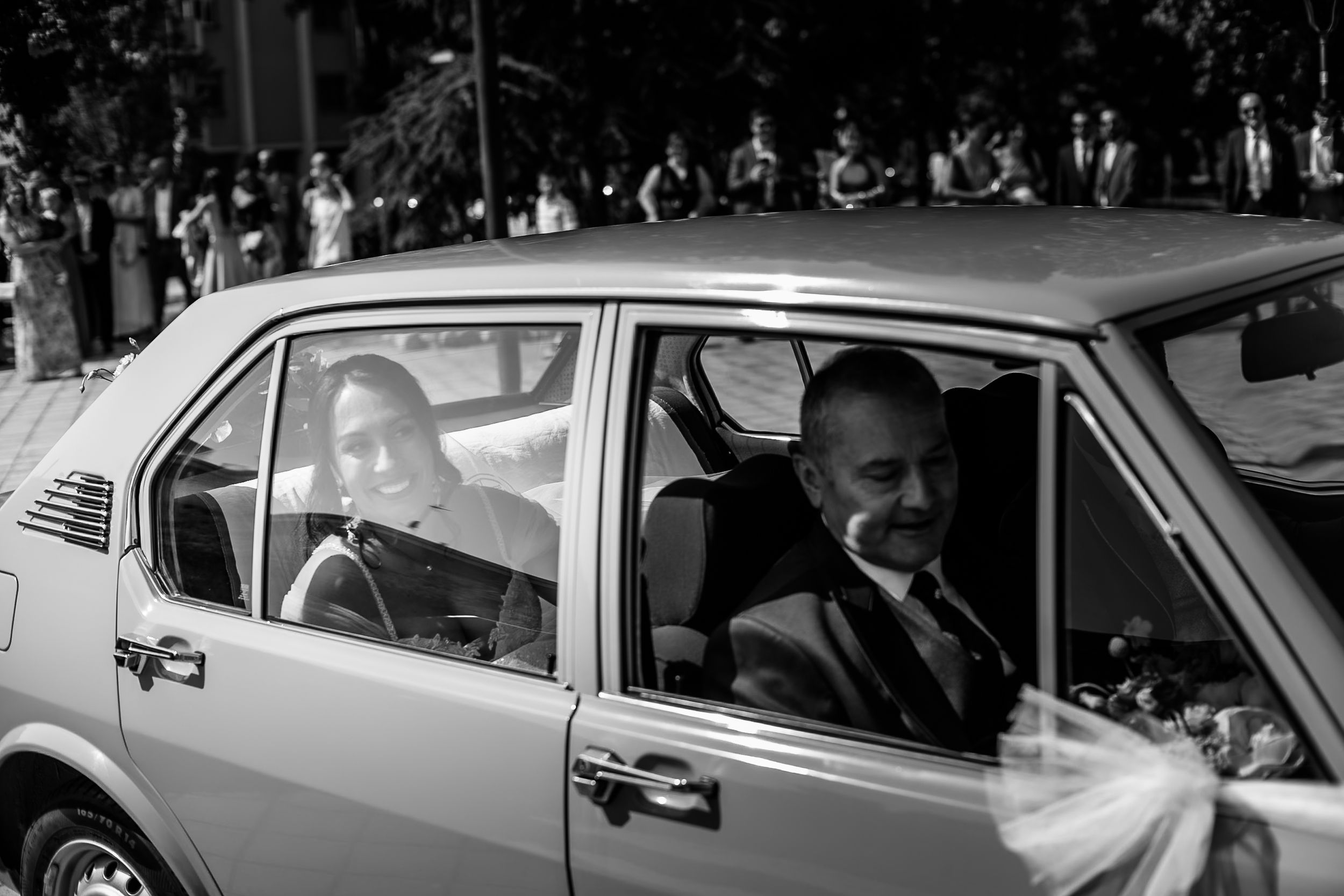 Black and white photo of a bride and groom smiling inside a vintage car decorated with white ribbons.