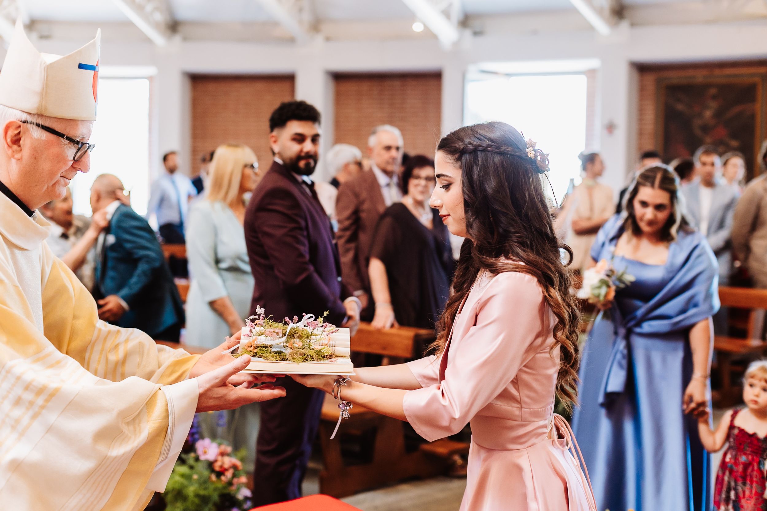 Priest offering a decorated tray to a woman in a pink dress during a church wedding ceremony.