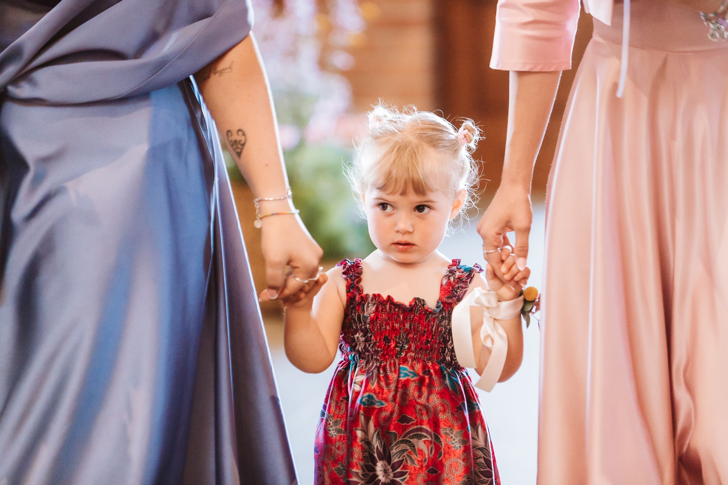 A toddler girl in a floral dress holds hands with two women in pastel gowns at a wedding ceremony.