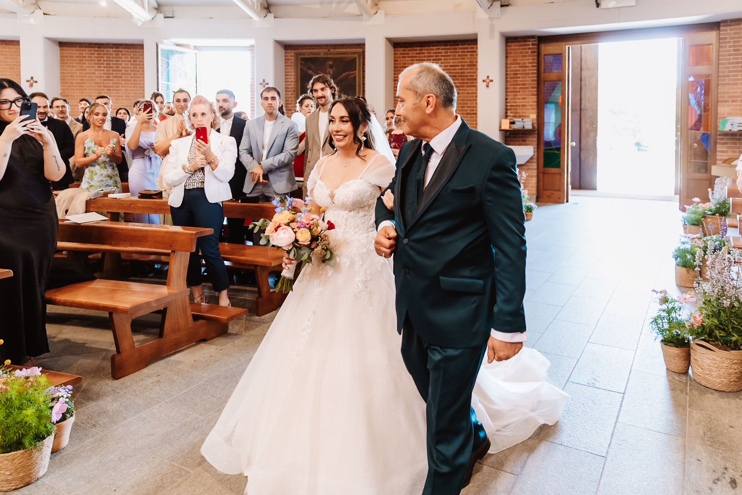 Bride in white gown walks down church aisle with older man, guests applauding, holding colorful bouquet.