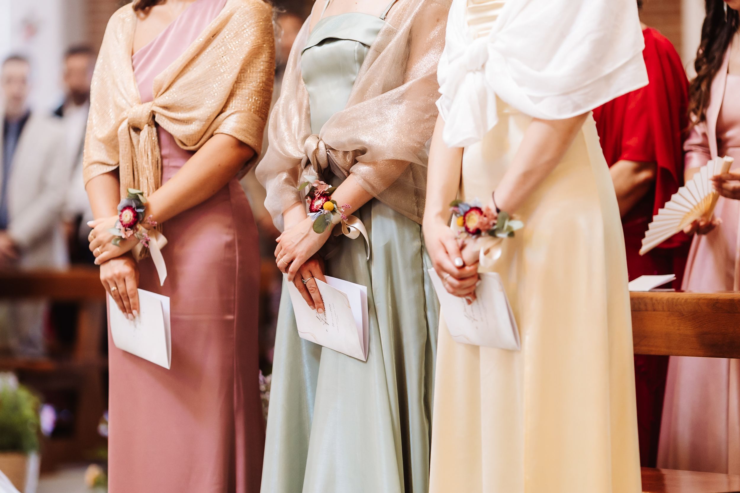 Bridesmaids in pastel dresses holding floral wrist corsages and programs during a wedding ceremony.