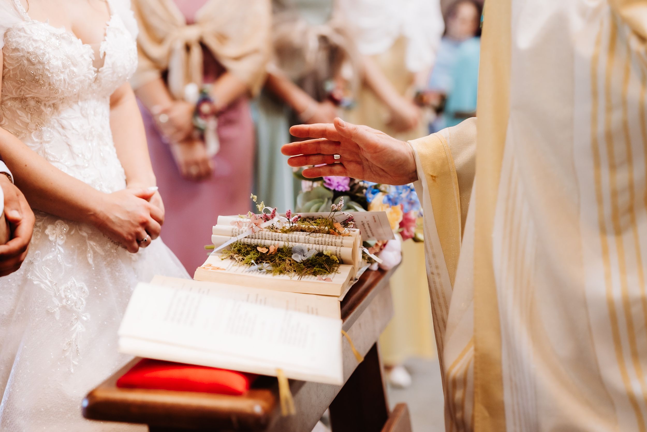 Priest blessing wedding rings on a Bible during a church ceremony with bride in white lace gown.