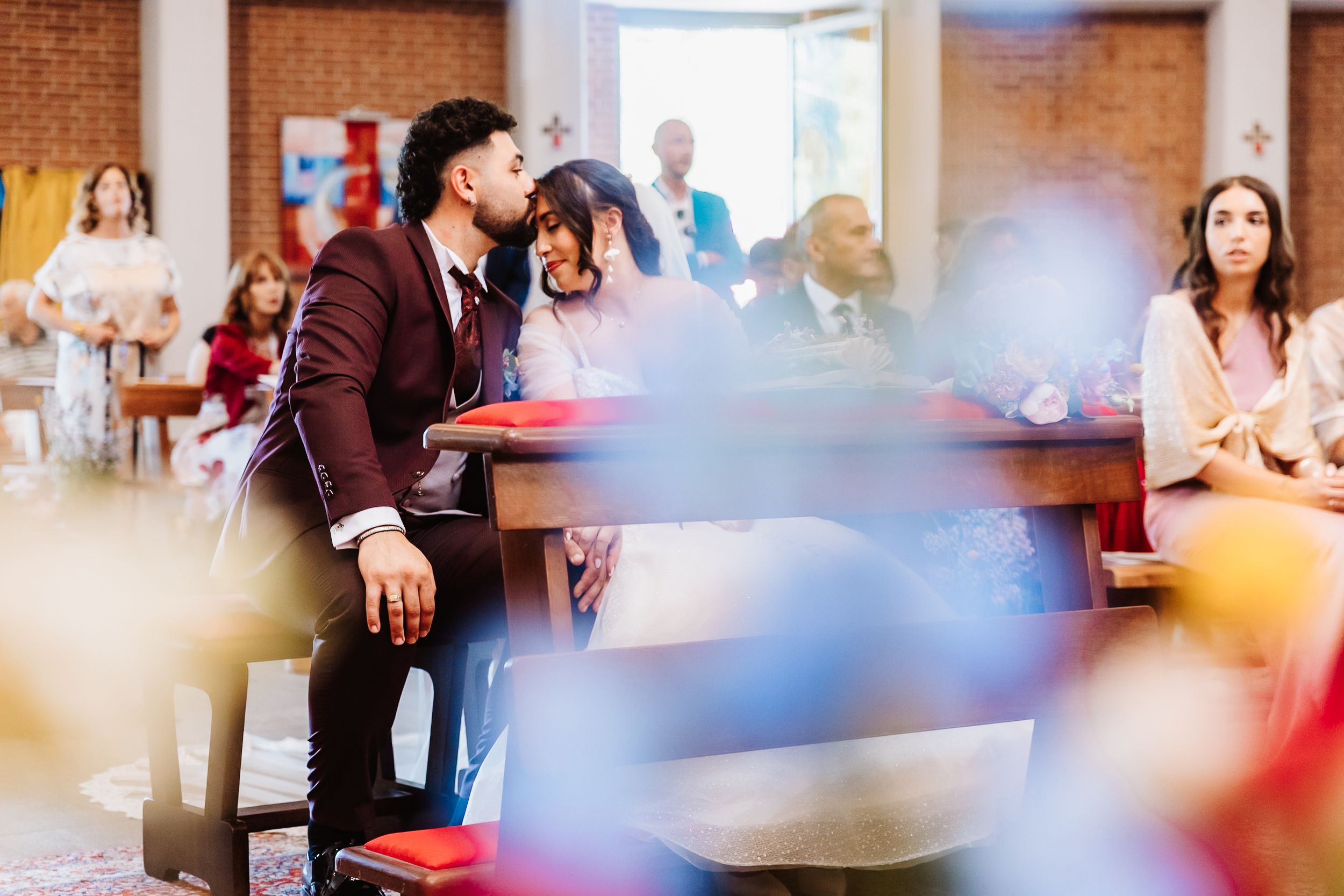 Couple sharing an intimate moment in church pews during a wedding ceremony, surrounded by guests in formal attire.