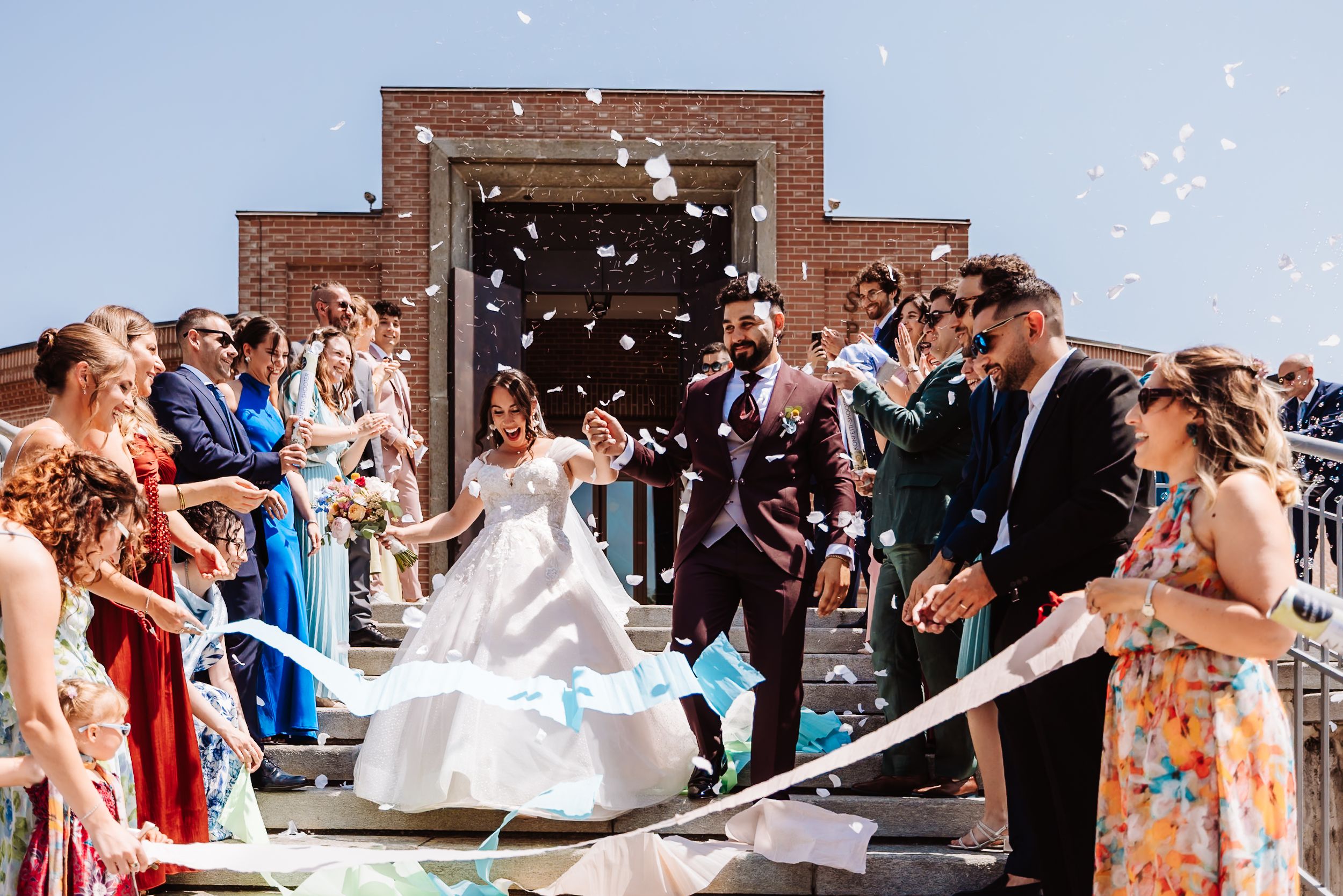 Bride and groom exit church amid confetti shower, surrounded by cheering guests in colorful attire on sunny wedding day.