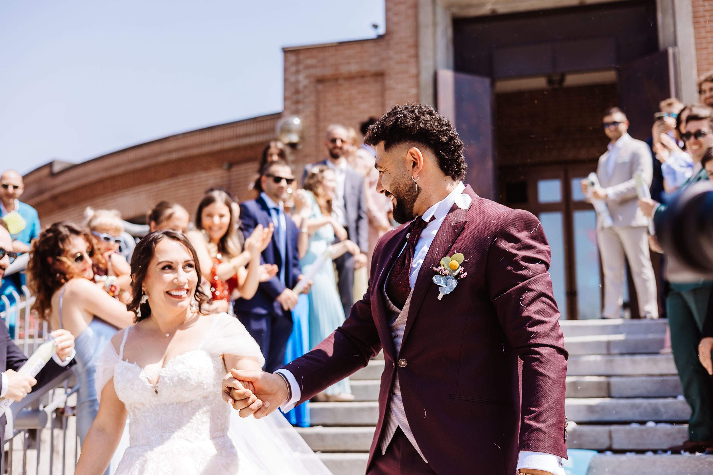 Bride and groom holding hands, smiling on church steps, surrounded by cheering guests at a sunny outdoor wedding ceremony.