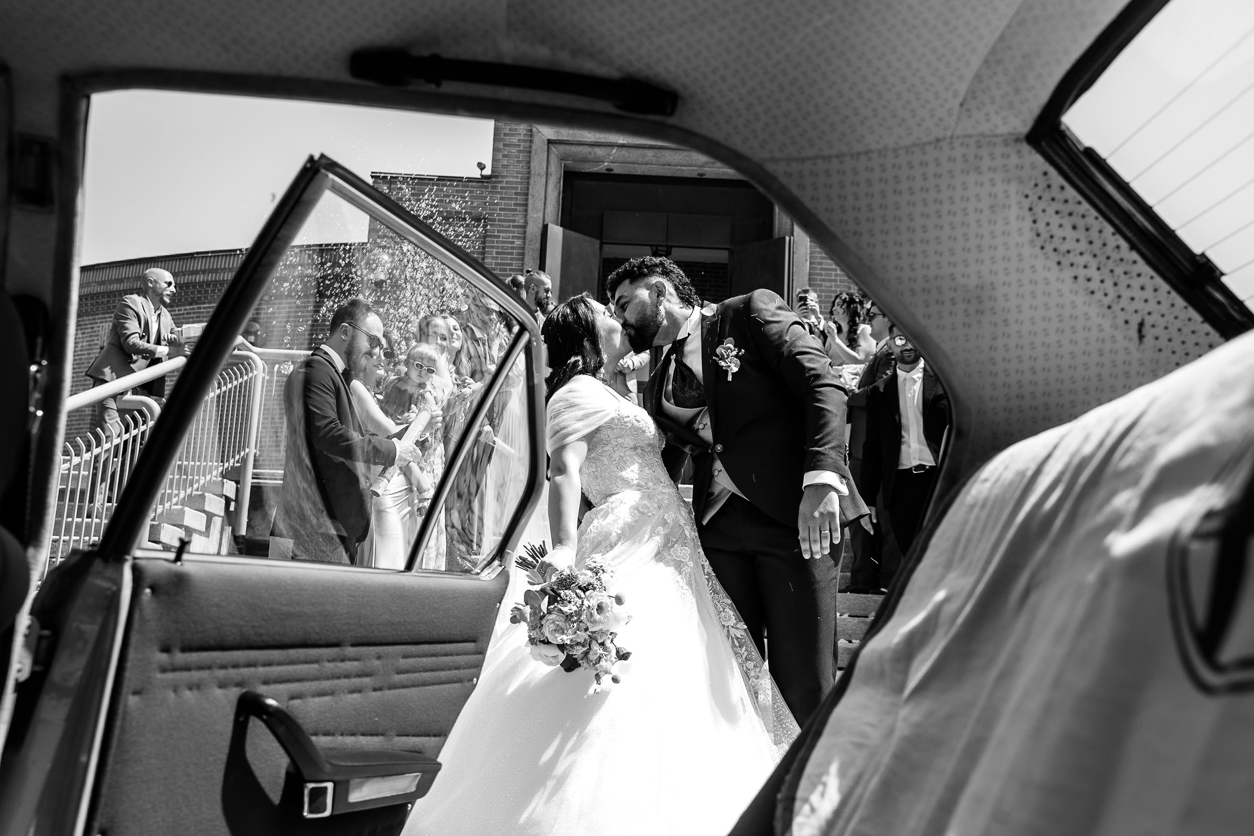 Black and white wedding photo of bride and groom kissing outside church, captured through vintage car door.