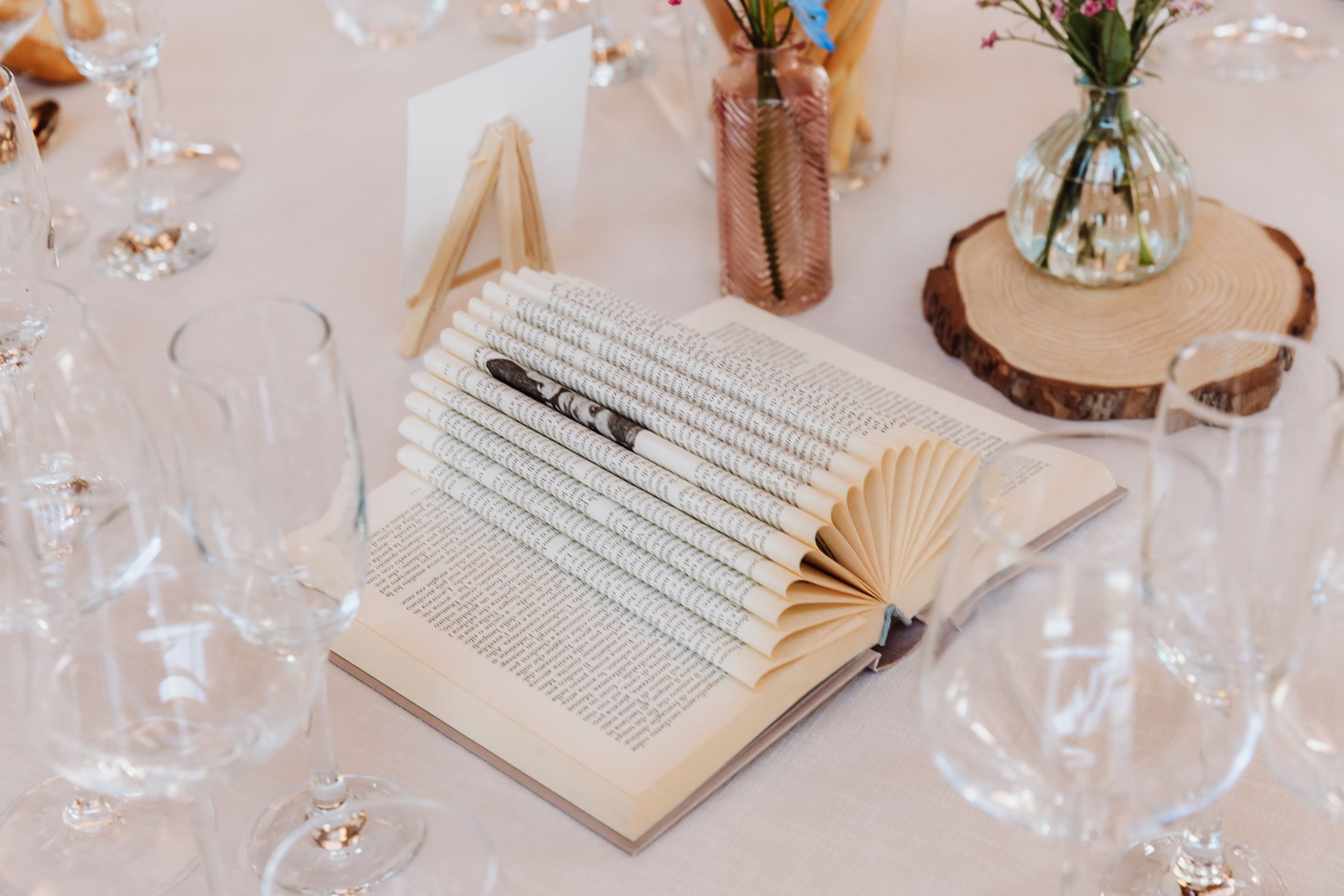 Elegant wedding table setting with an open book centerpiece, folded pages, pencil, glasses, and floral decor on a wood slice.
