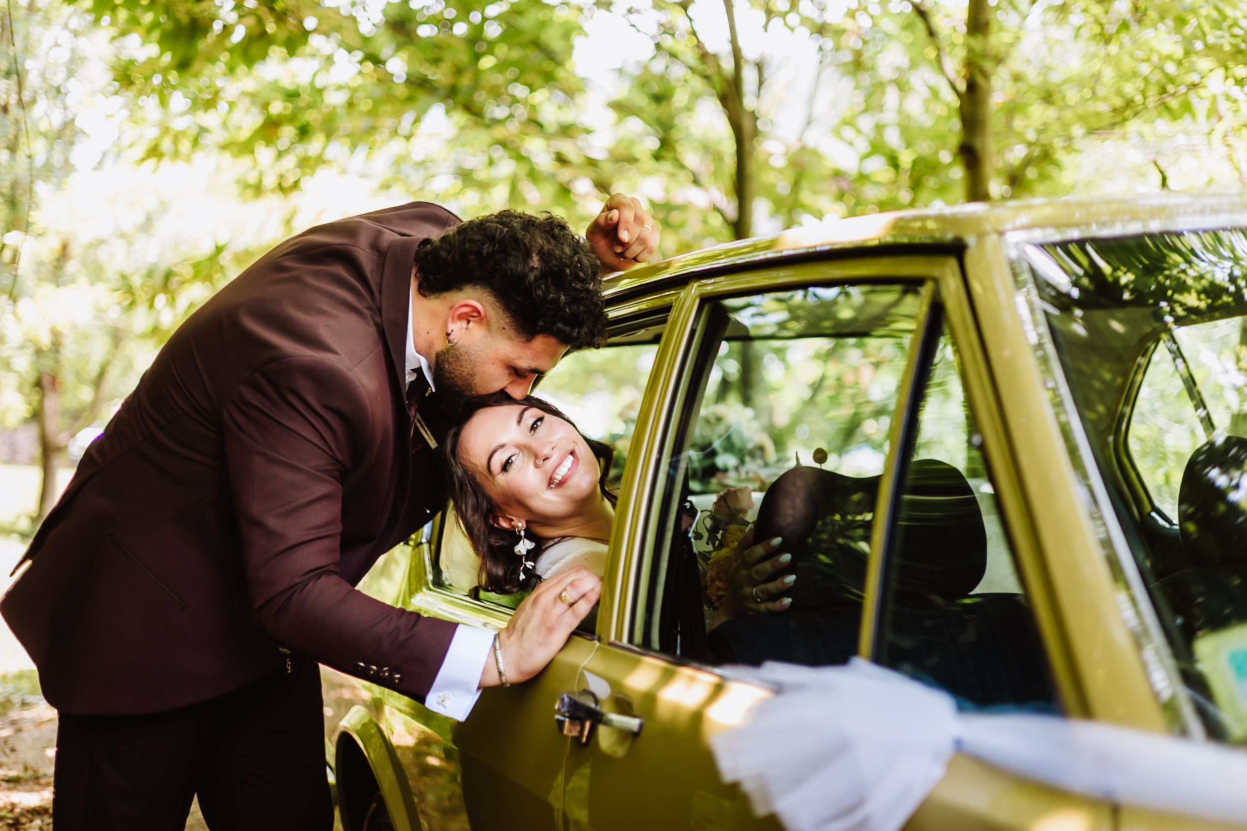 Groom in burgundy suit kissing bride through yellow vintage car window outdoors.