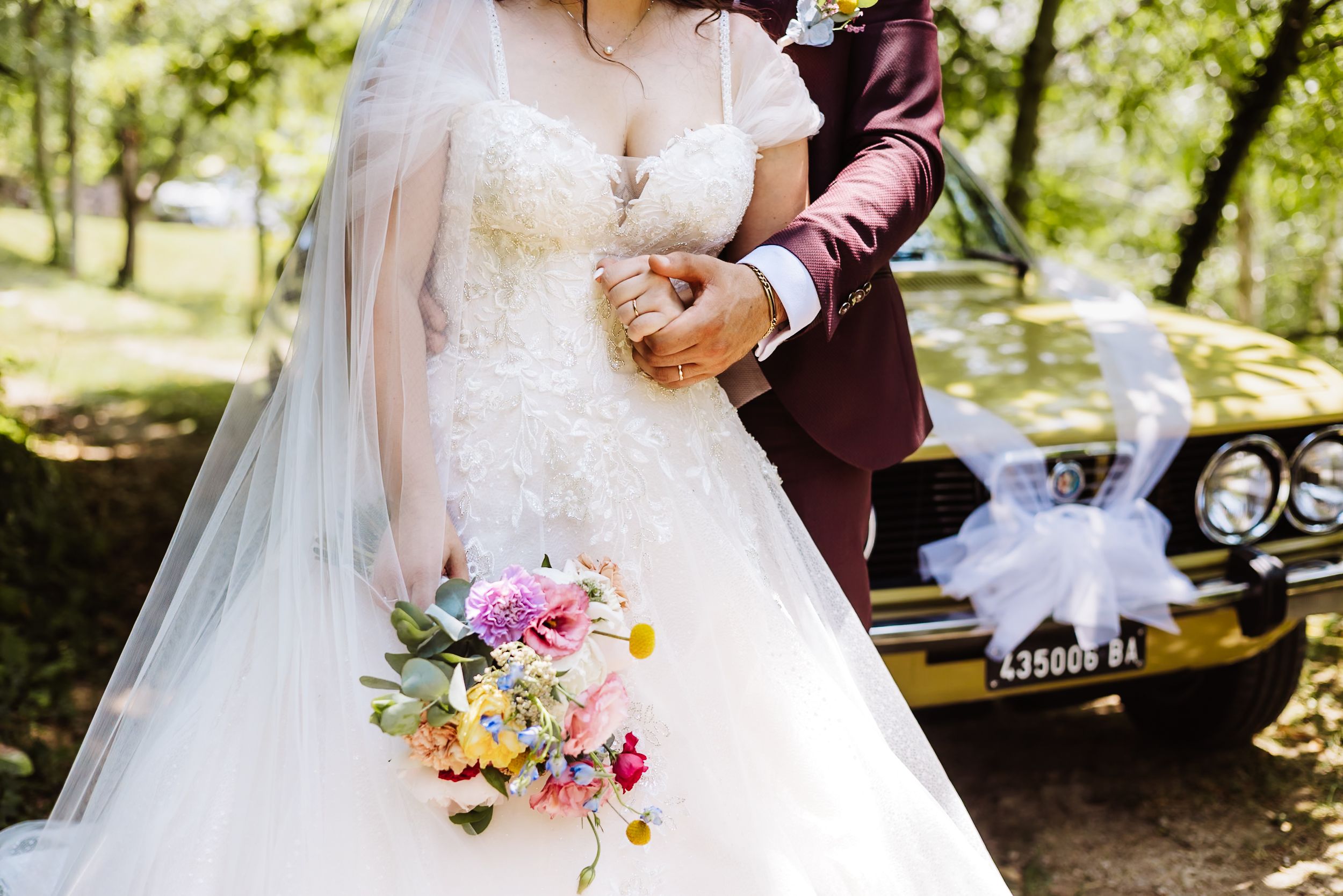 Bride in lace gown holding colorful bouquet stands with groom in burgundy suit beside vintage yellow wedding car.