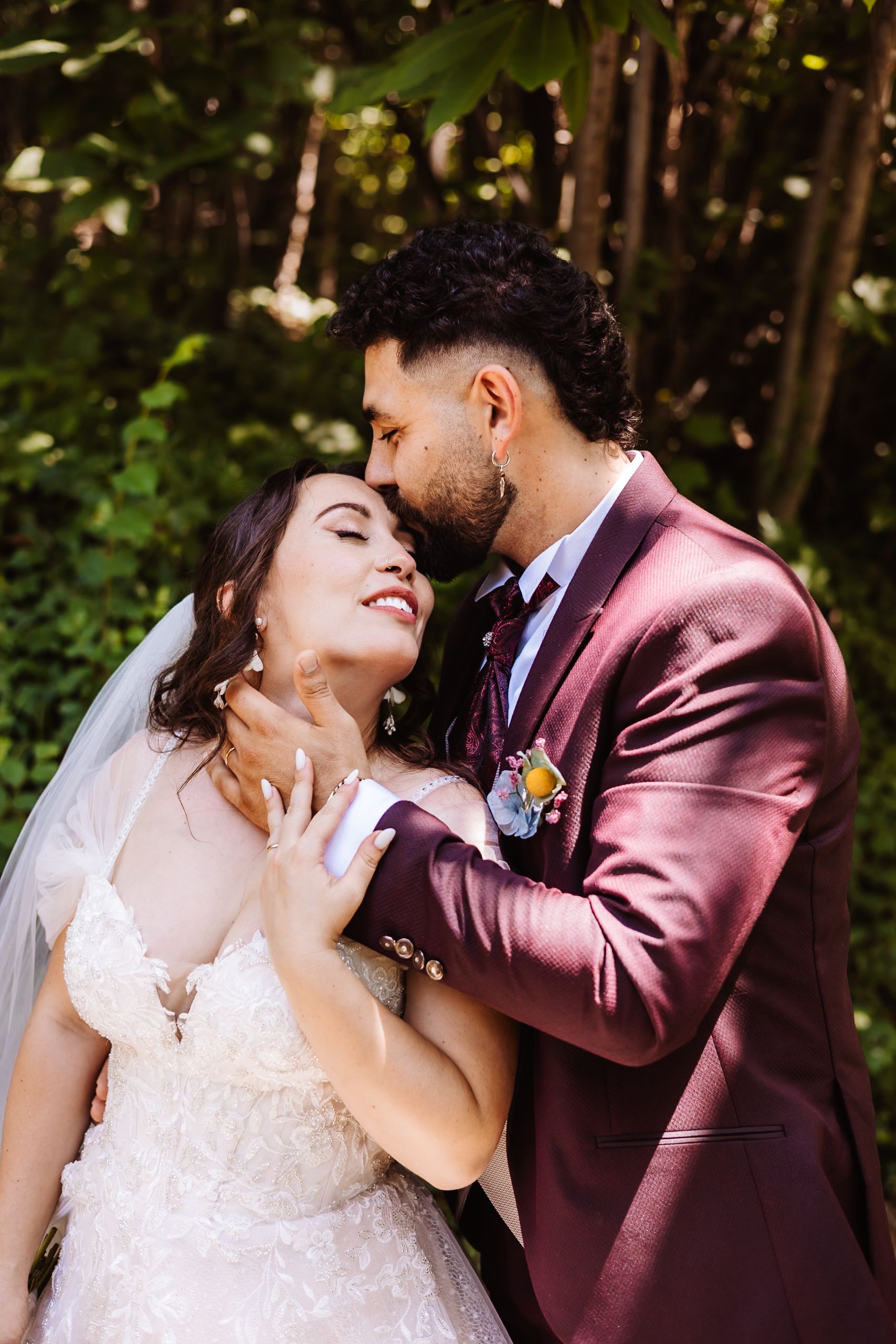 Bride in lace gown and groom in burgundy suit share romantic moment in lush garden.