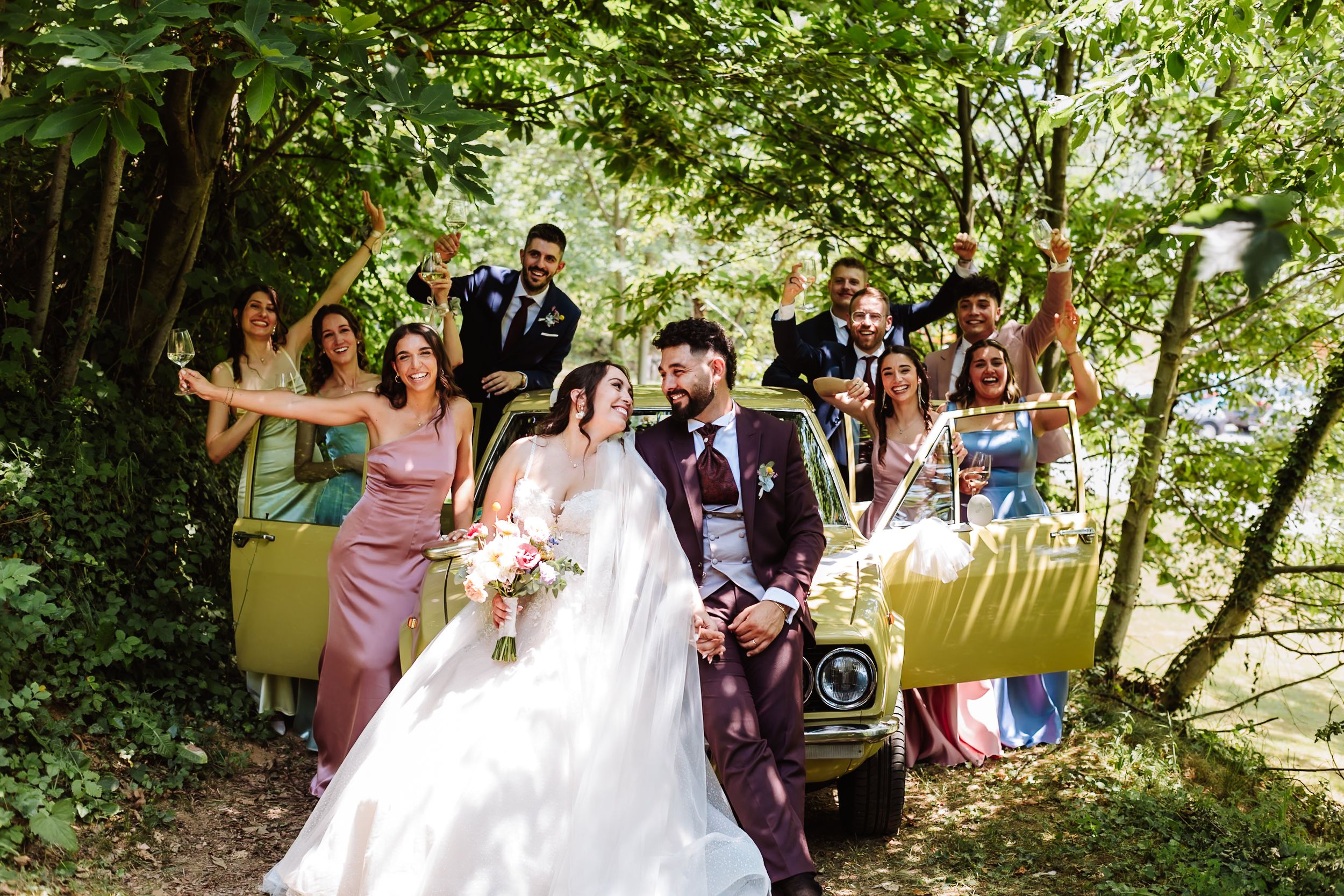 Wedding couple poses with bridal party near vintage car surrounded by lush green trees.