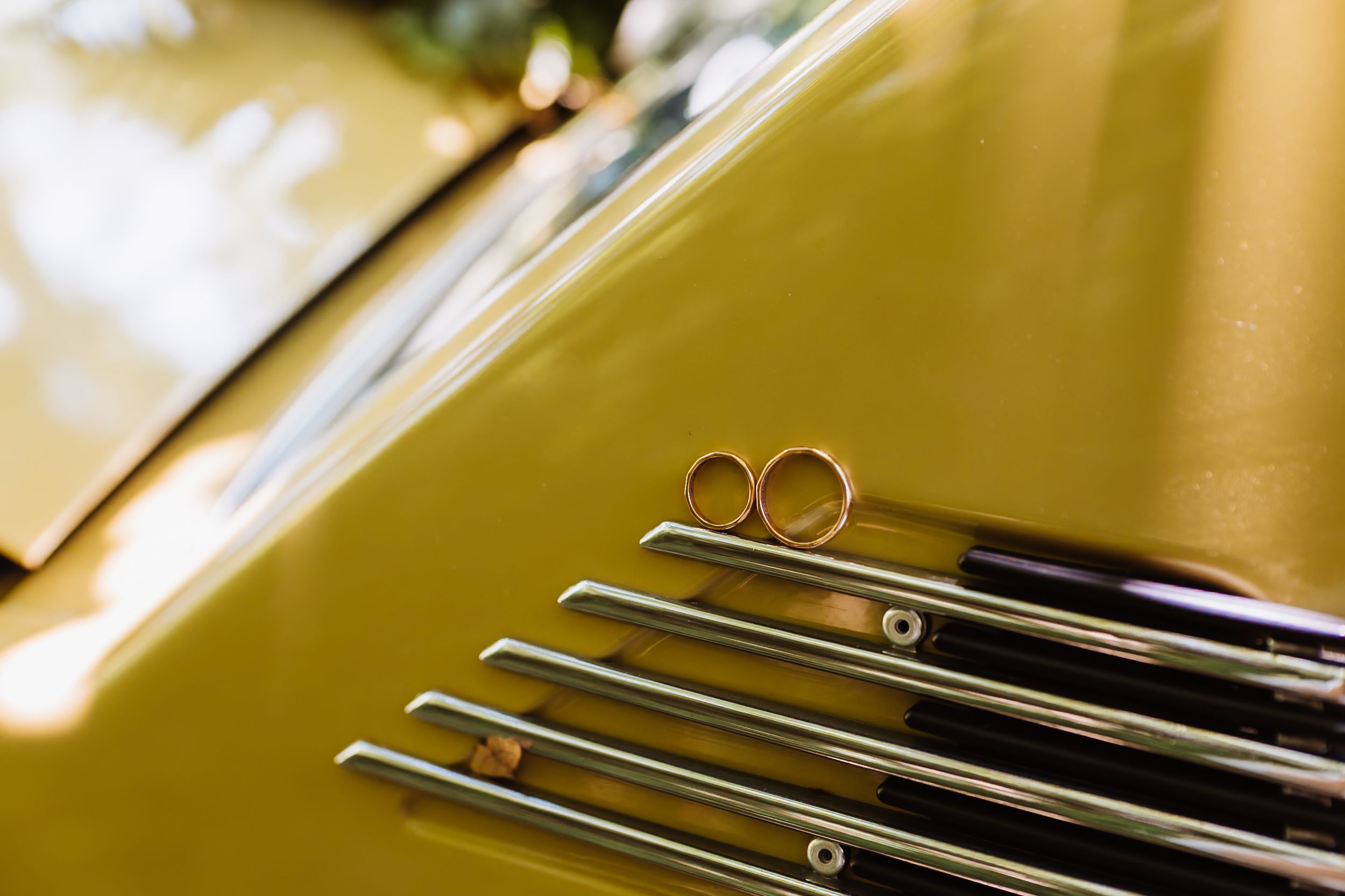 Two gold wedding rings resting on chrome grille of a vintage yellow classic car at wedding.