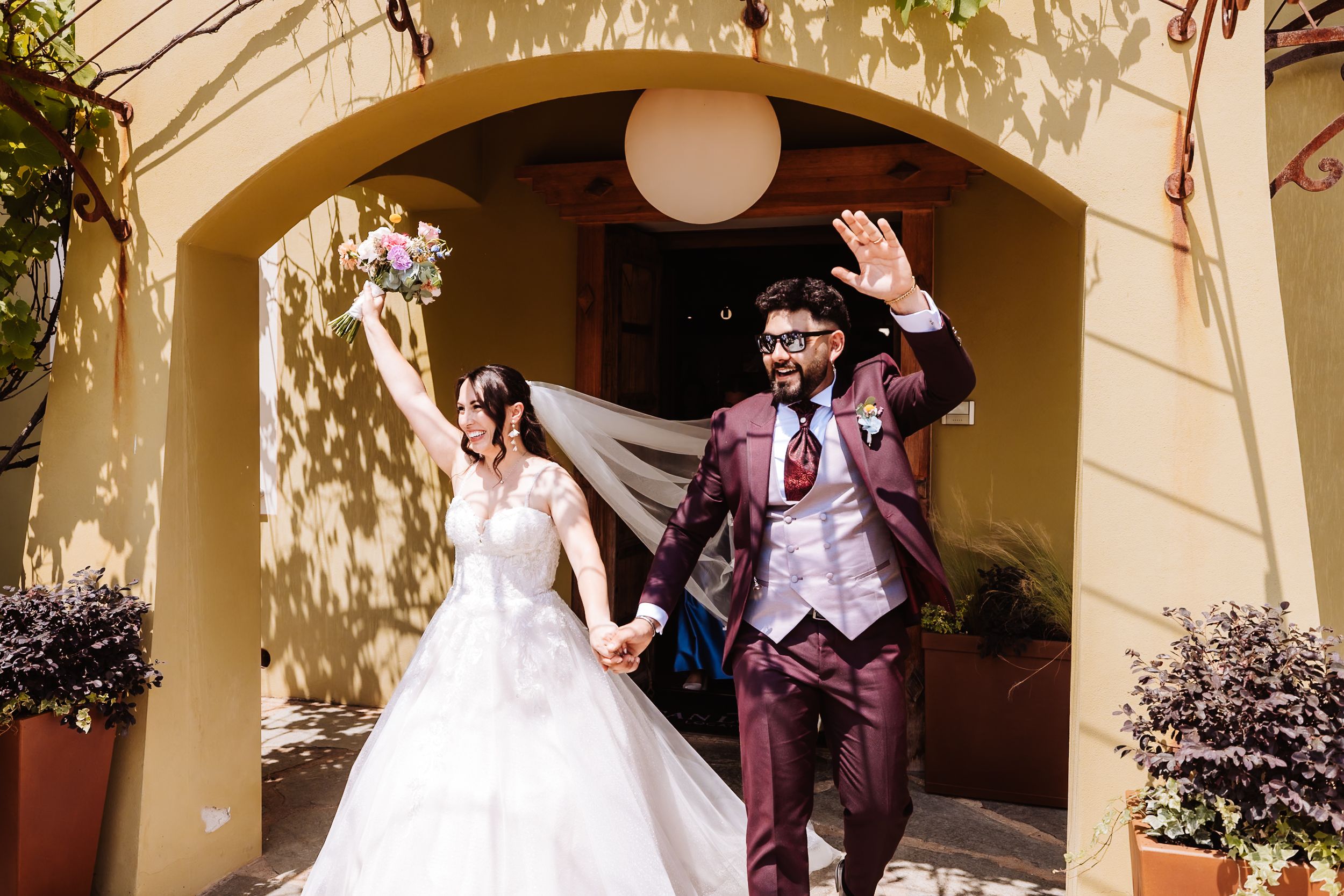 Joyful newlyweds exit venue waving, bride in white gown with veil, groom in burgundy suit.