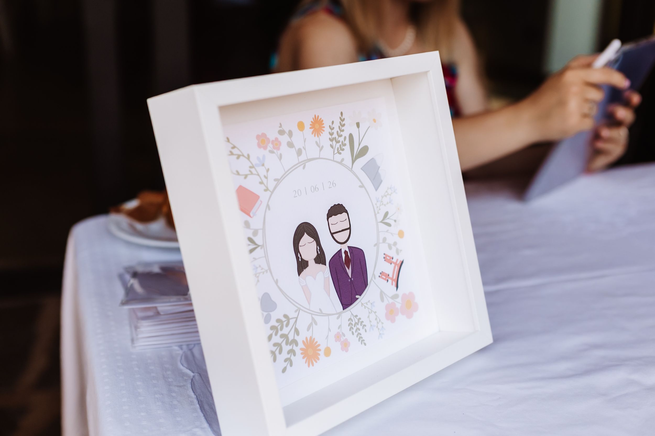 White framed illustrated portrait of a couple surrounded by floral wreath design, displayed on a table at a wedding reception.