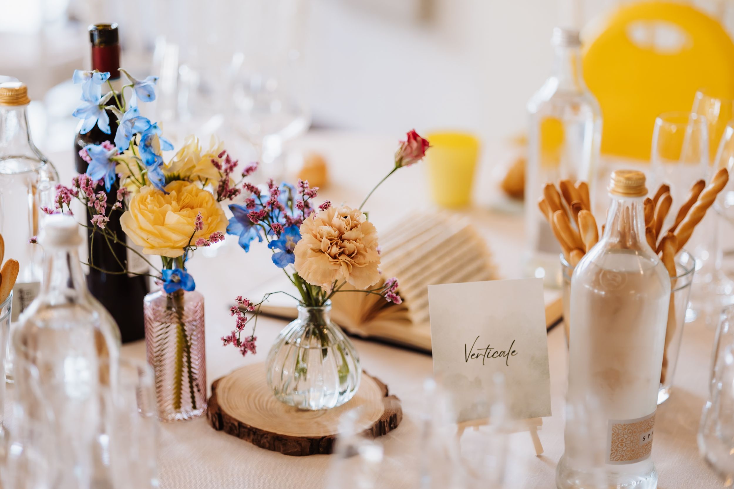 Elegant wedding table centerpiece with colorful wildflowers in glass vases on wood slice, bottles and glassware.