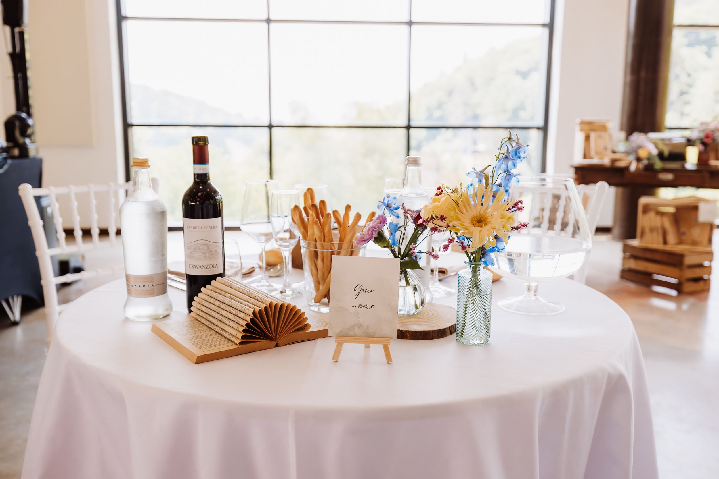 Elegant wedding reception table with wine bottle, breadsticks, wildflower centerpiece, and wooden fan on white tablecloth.