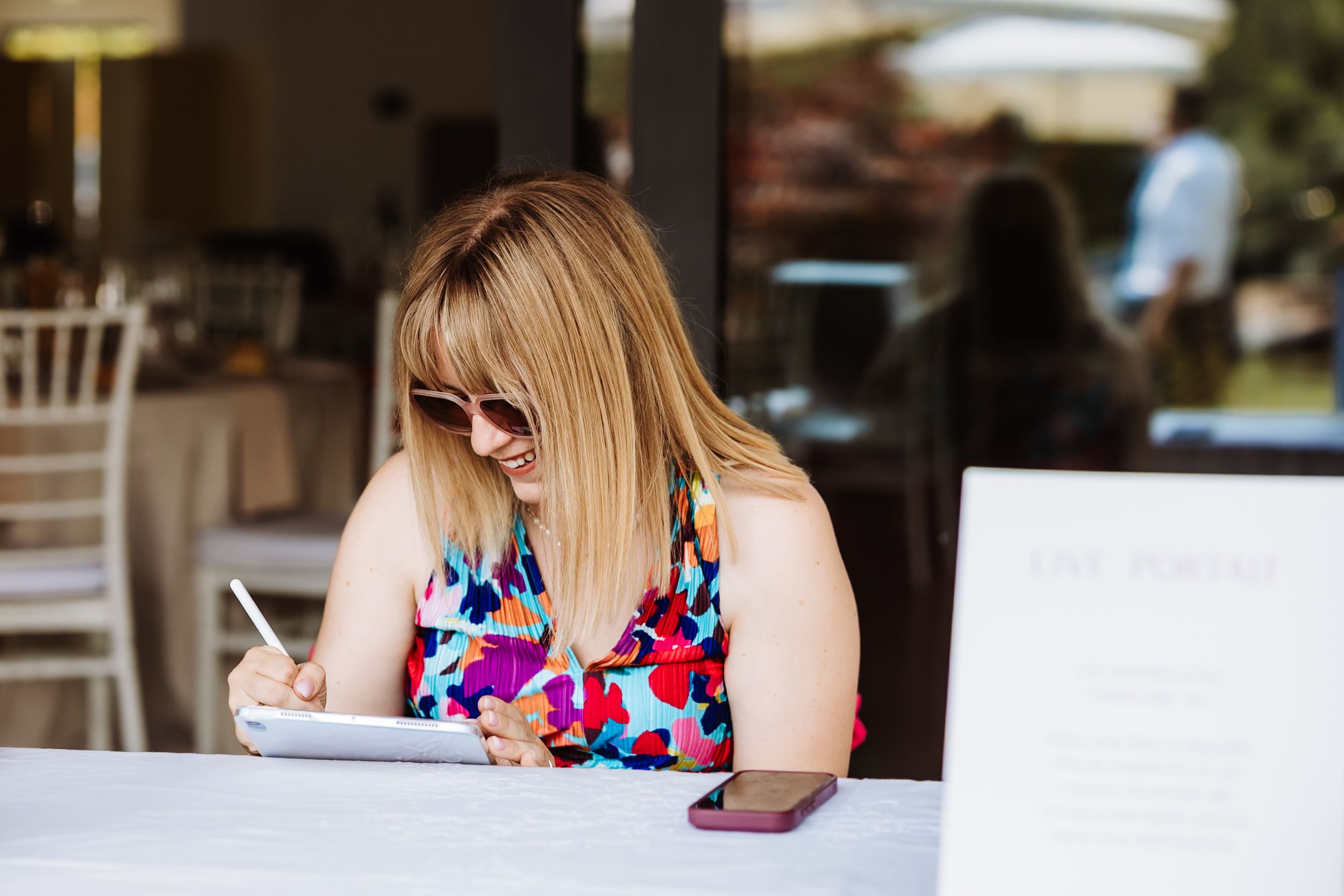 Blonde woman in floral dress writing notes at outdoor wedding venue table with sunglasses.