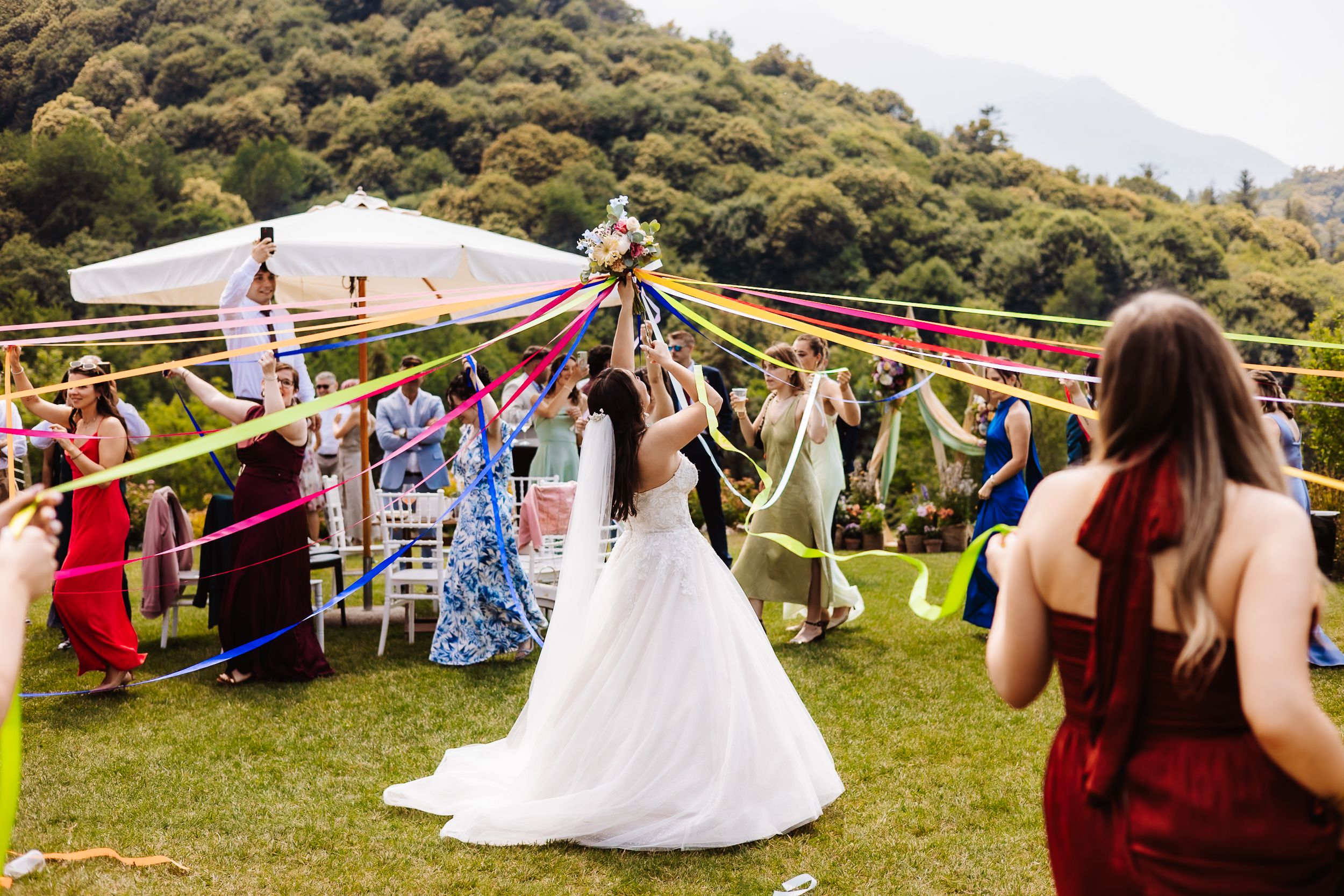 Bride in white gown plays maypole with colorful ribbons surrounded by guests at outdoor wedding celebration.