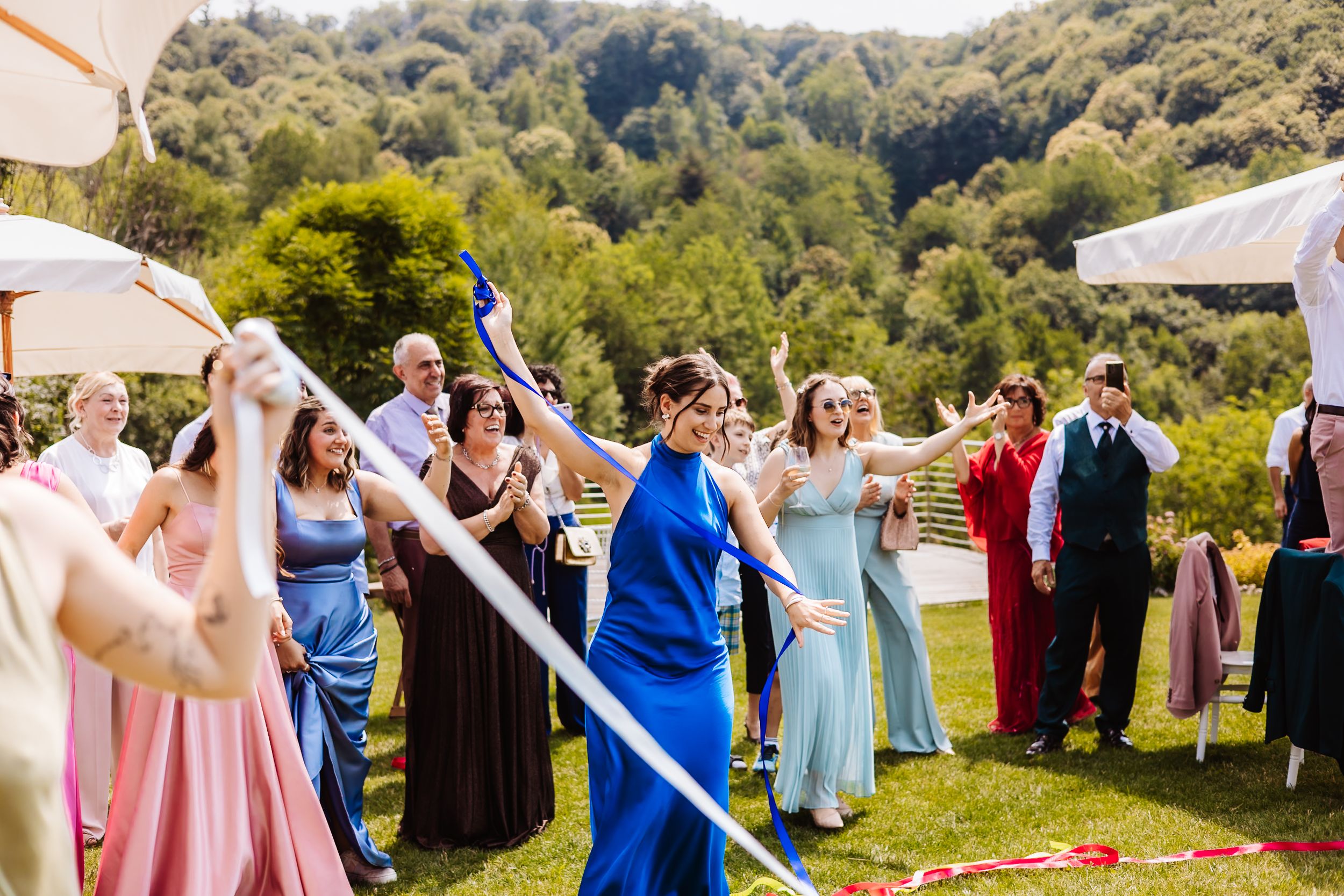 Bridesmaids in colorful dresses dancing and celebrating outdoors at a wedding reception with lush green hills.