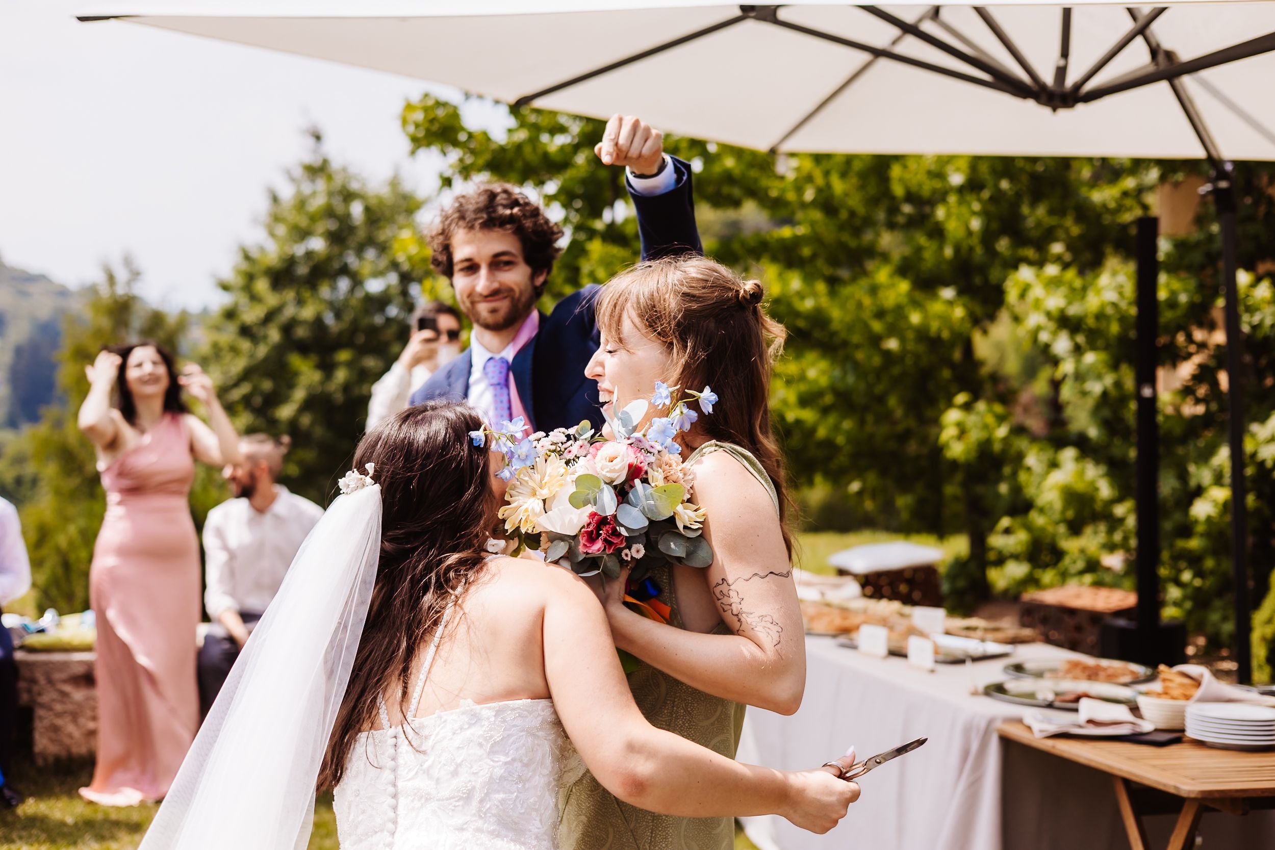 Bride hugging bridesmaid outdoors at wedding reception while groom celebrates in background.