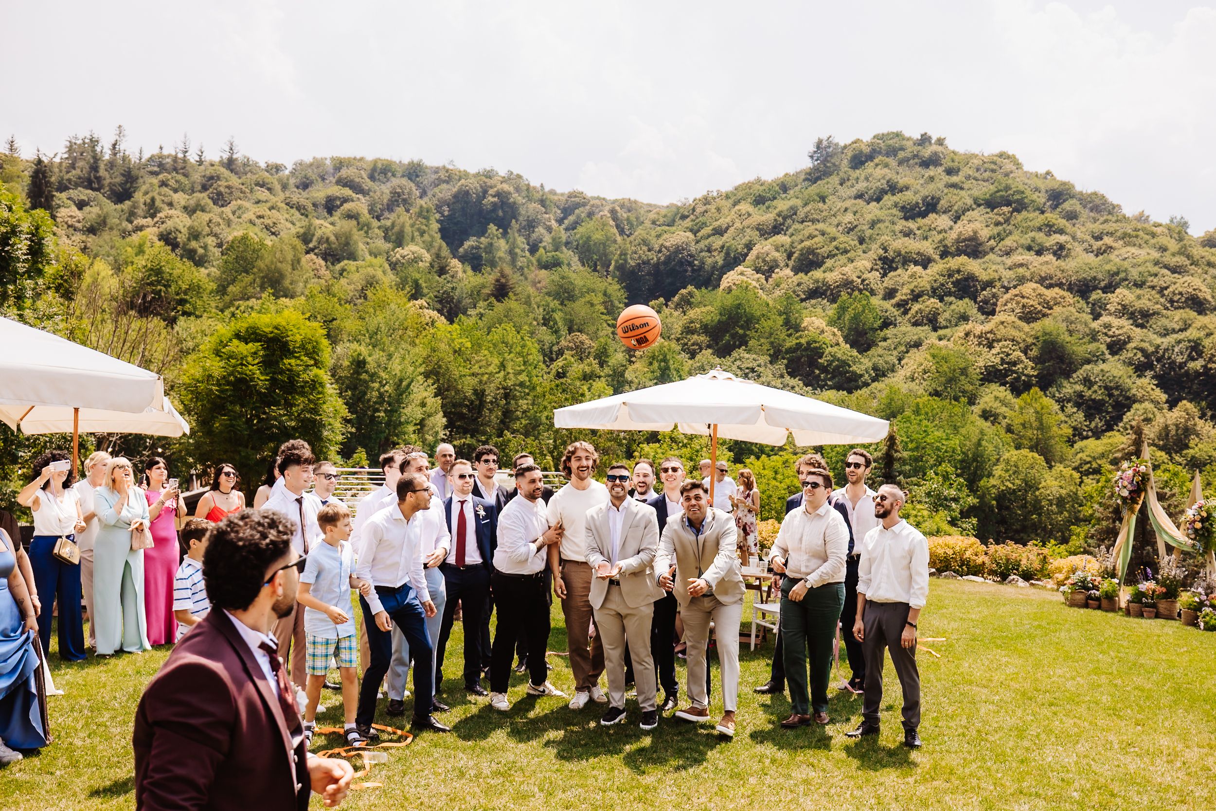 Groomsmen tossing a bouquet at an outdoor wedding reception with lush green hills in the background.