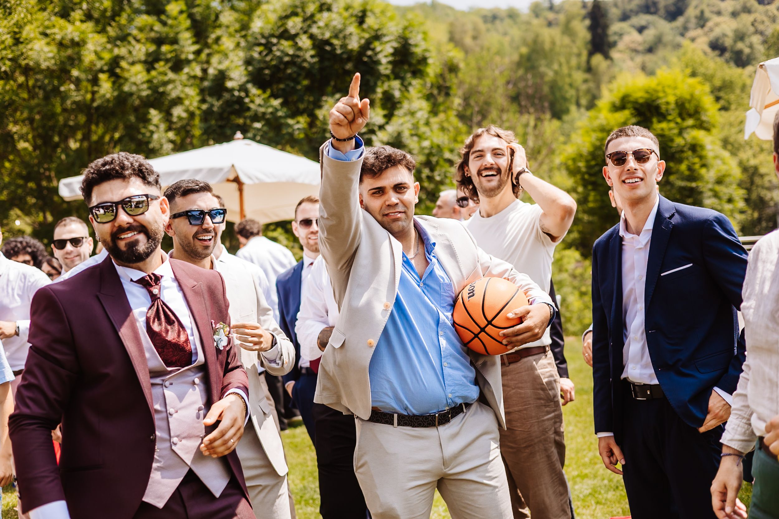 Groomsmen in suits celebrating outdoors at a wedding, one holding a basketball while pointing skyward.