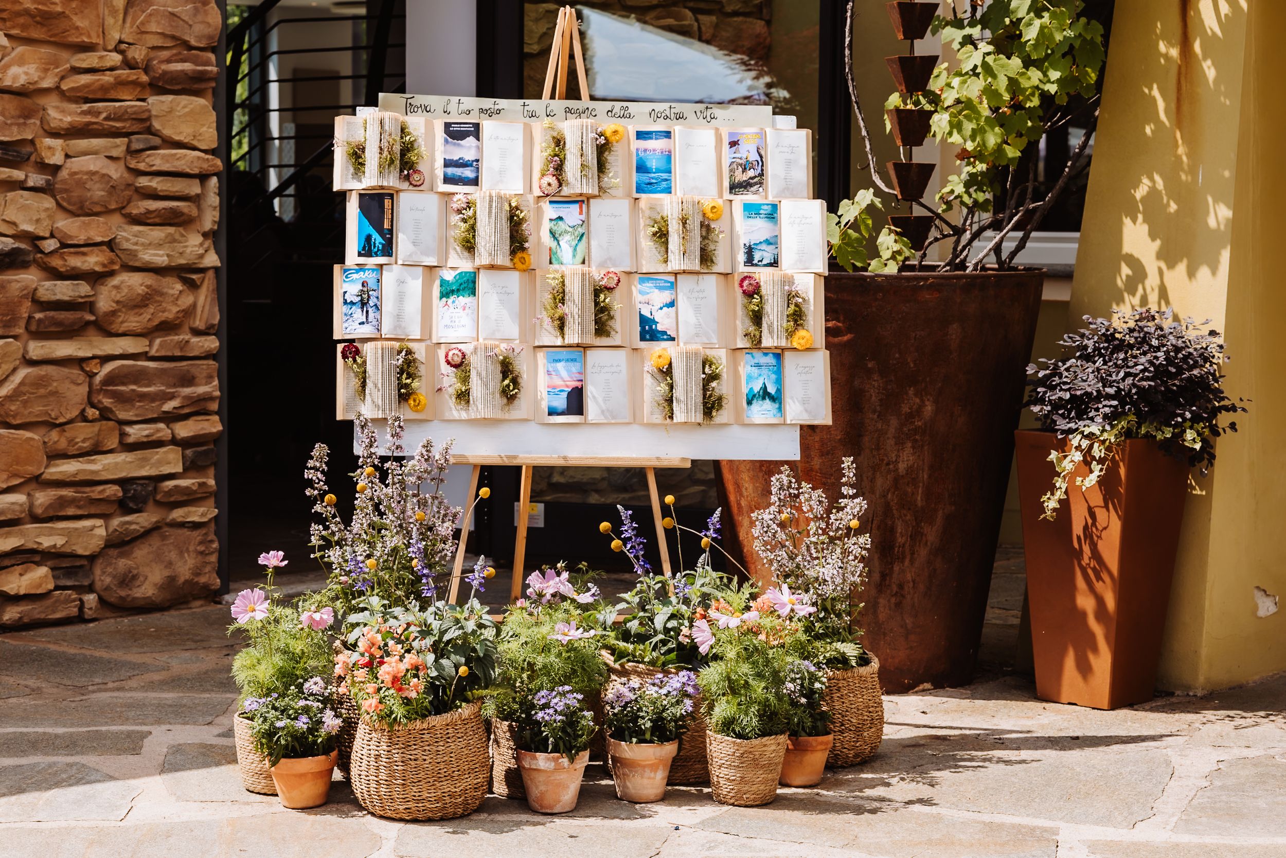 Elegant wedding seating chart display on easel surrounded by colorful potted flowers at outdoor venue.