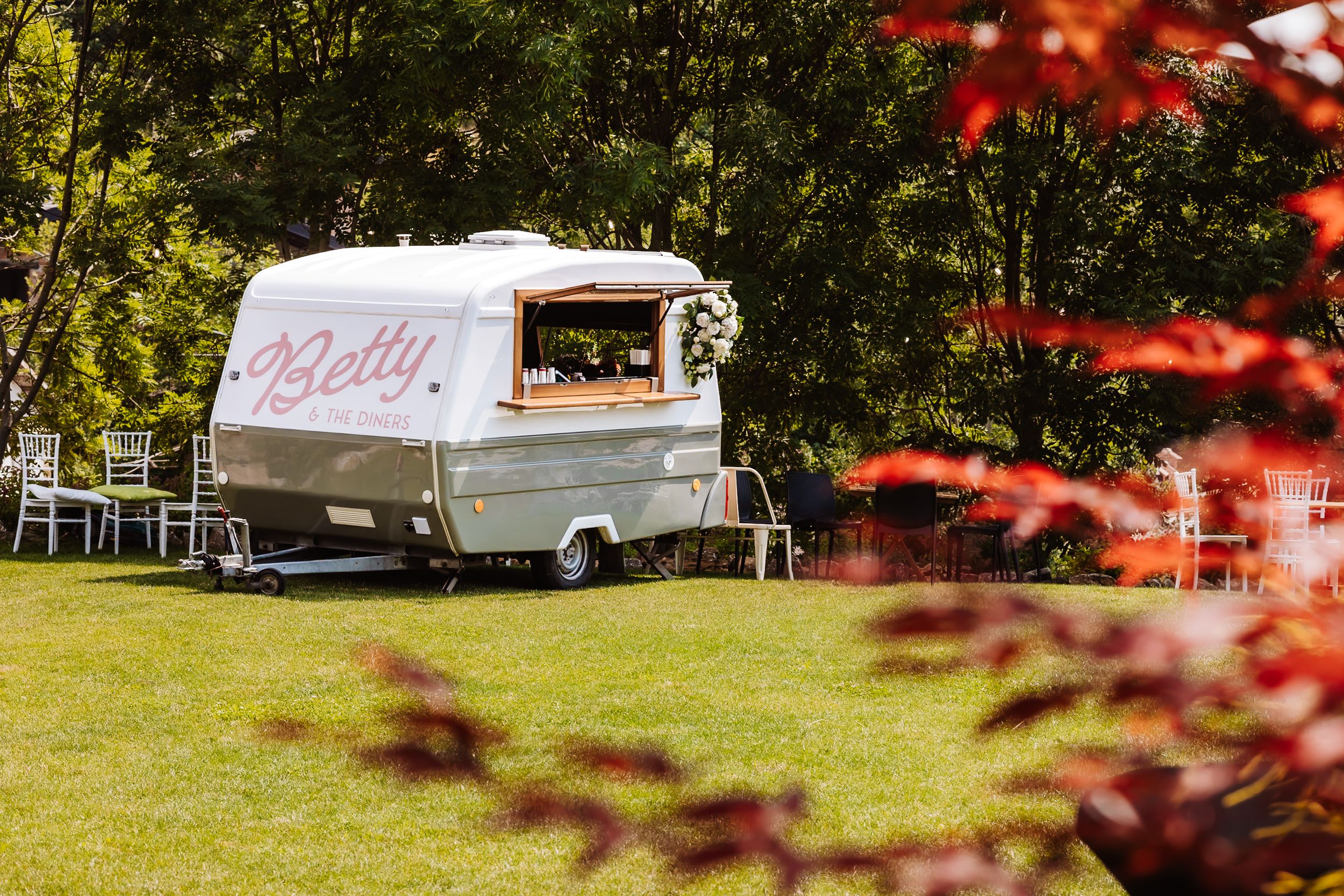 Vintage silver Betty caravan bar decorated with flowers parked on green lawn at outdoor wedding venue.