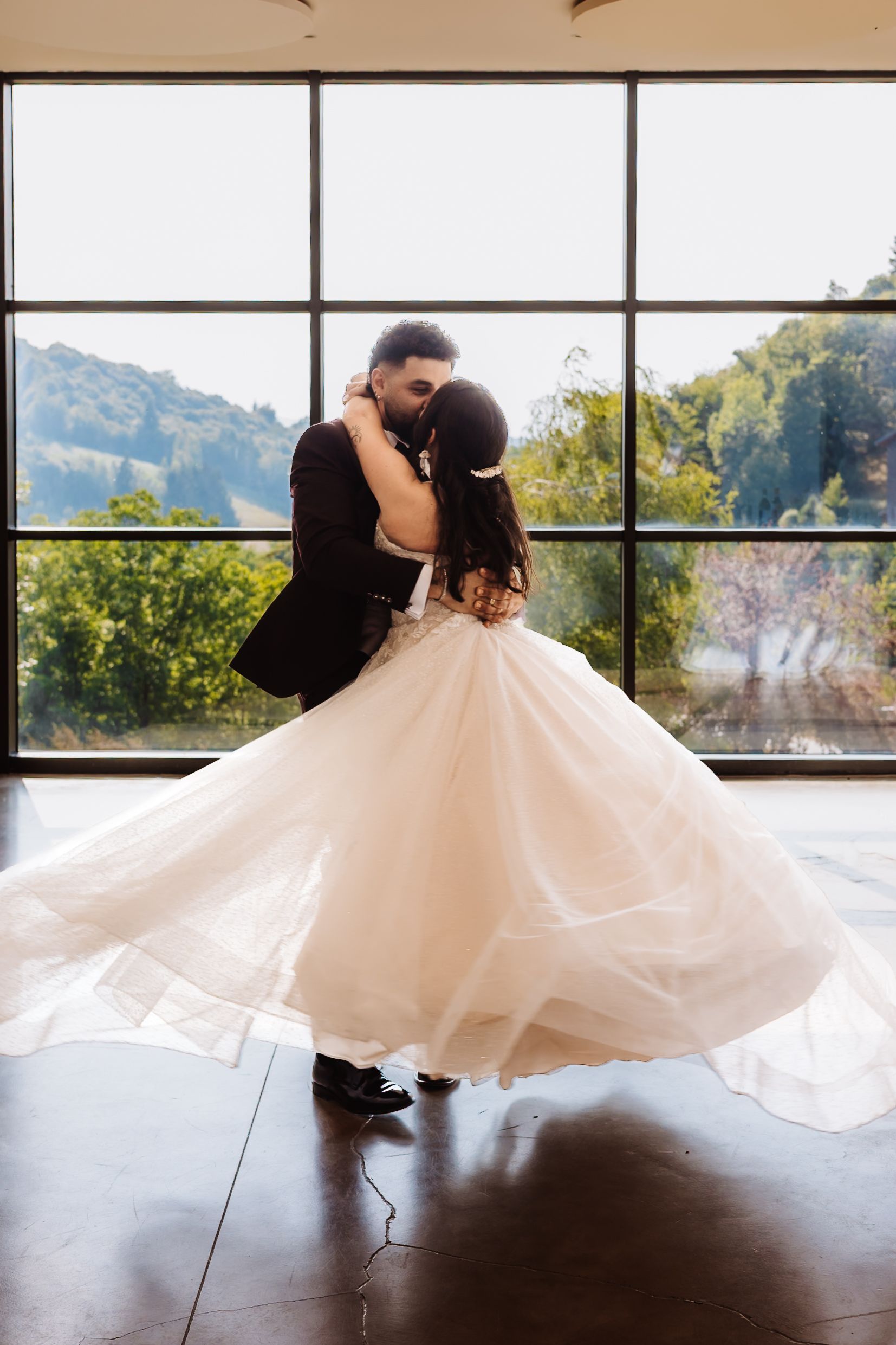 Bride and groom sharing their first dance, her white ballgown twirling before a large window with nature views.