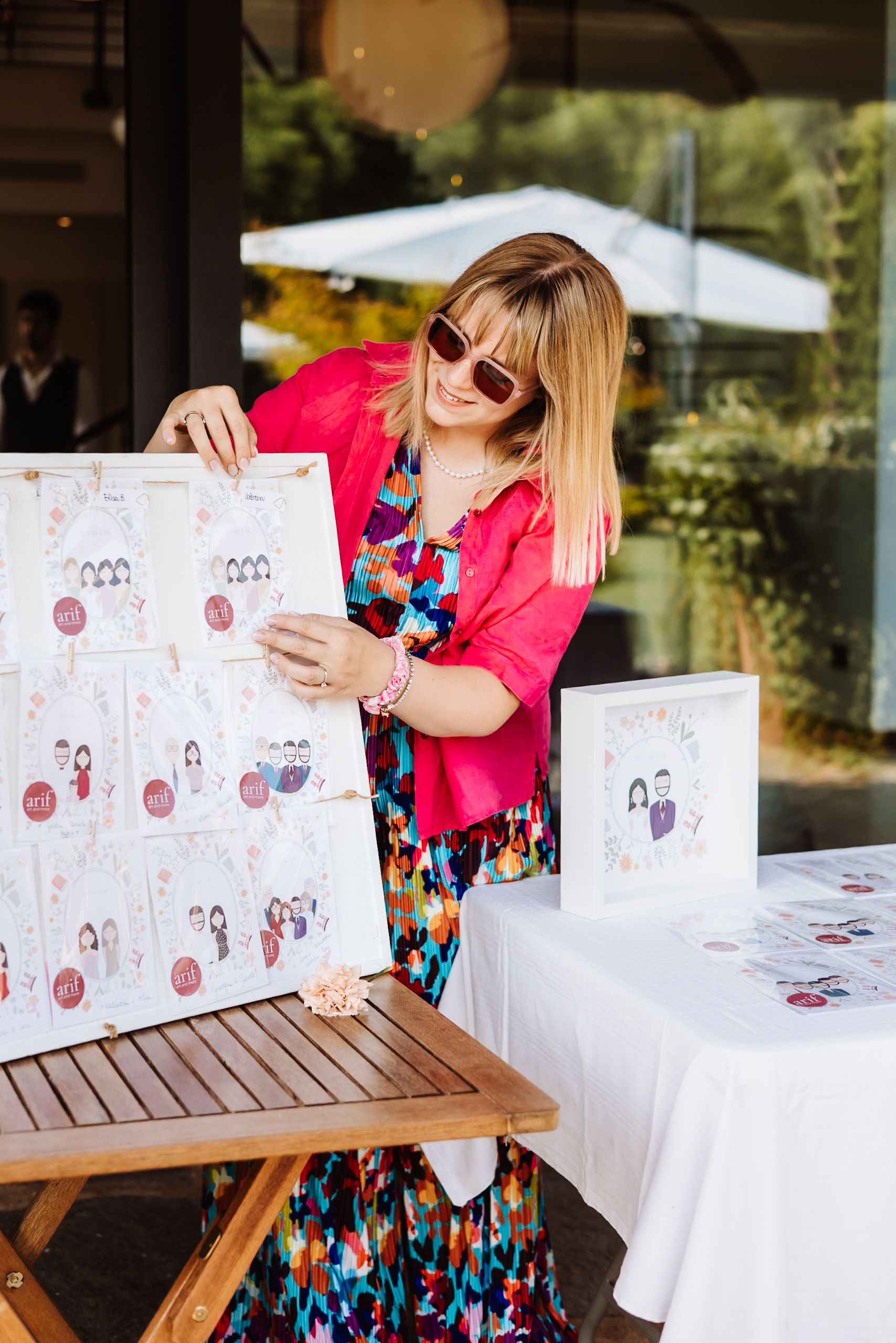 Woman in floral dress and pink cardigan displaying illustrated seating chart at outdoor wedding reception table.
