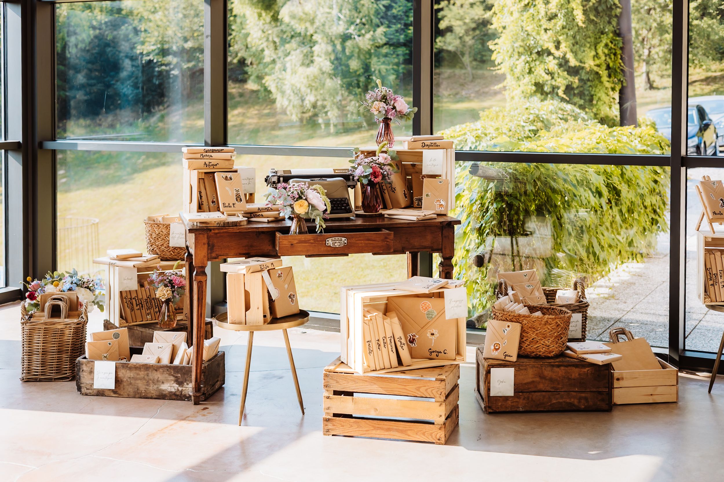 Rustic wedding display with wooden crates, wicker baskets, and vintage desk near large windows overlooking lush greenery.