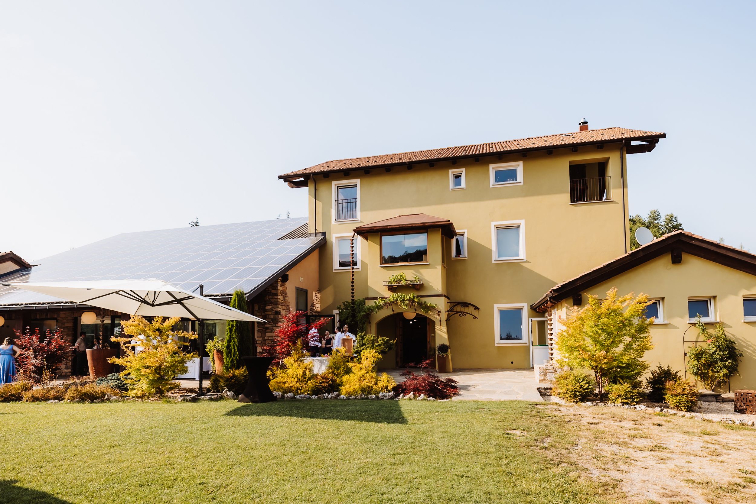 Yellow two-story house with solar panels, lush garden, colorful flowers, and green lawn on a sunny day.
