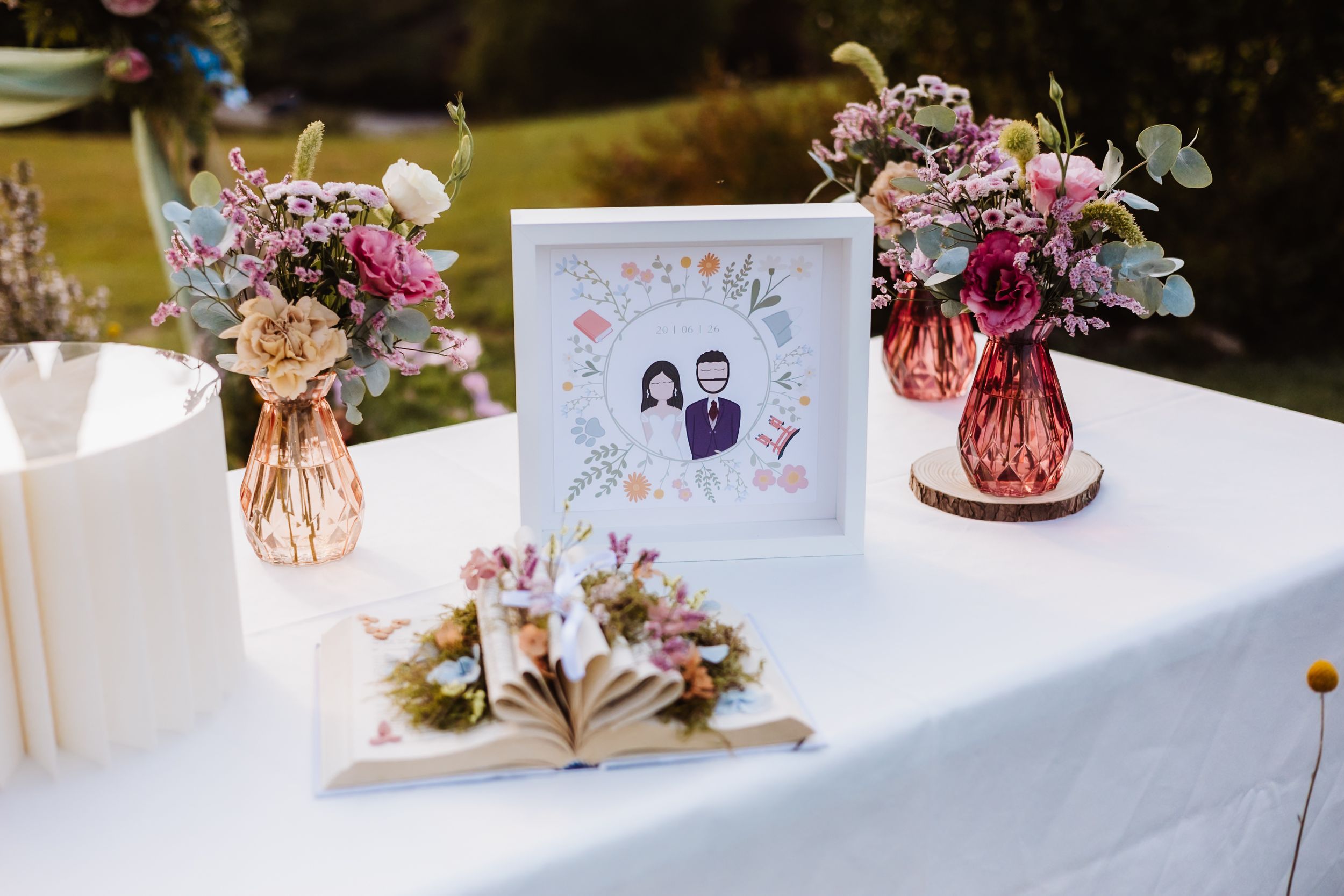Wedding table with floral arrangements, framed couple portrait, and open book centerpiece on white tablecloth outdoors.