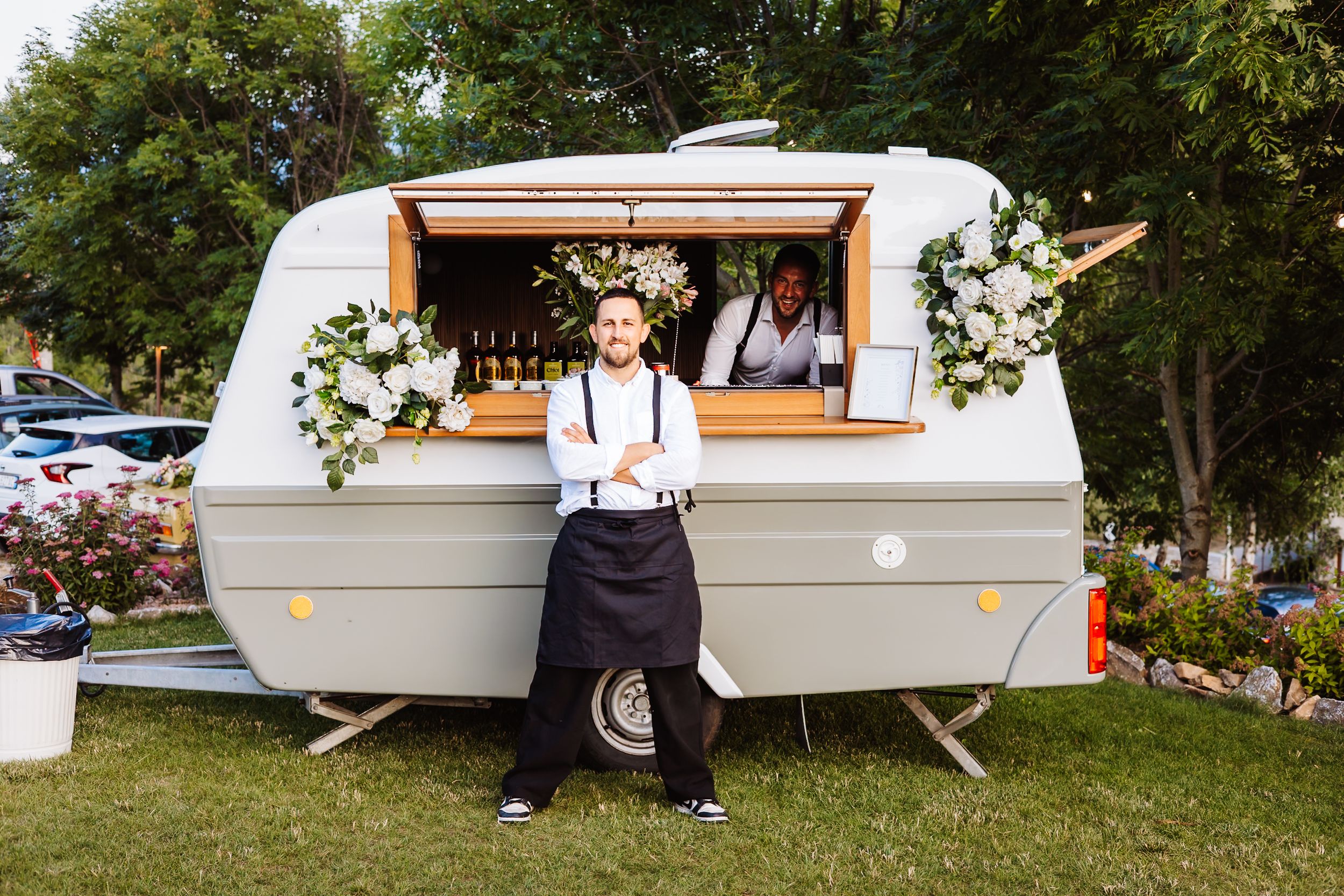 A smiling chef stands before a decorated food truck caravan adorned with white floral arrangements at an outdoor event.