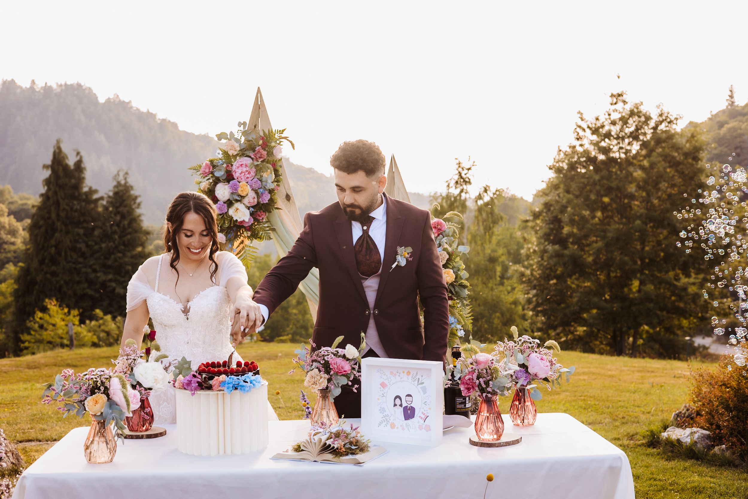 Bride and groom cutting wedding cake outdoors on a floral-decorated table with a scenic mountain church backdrop.