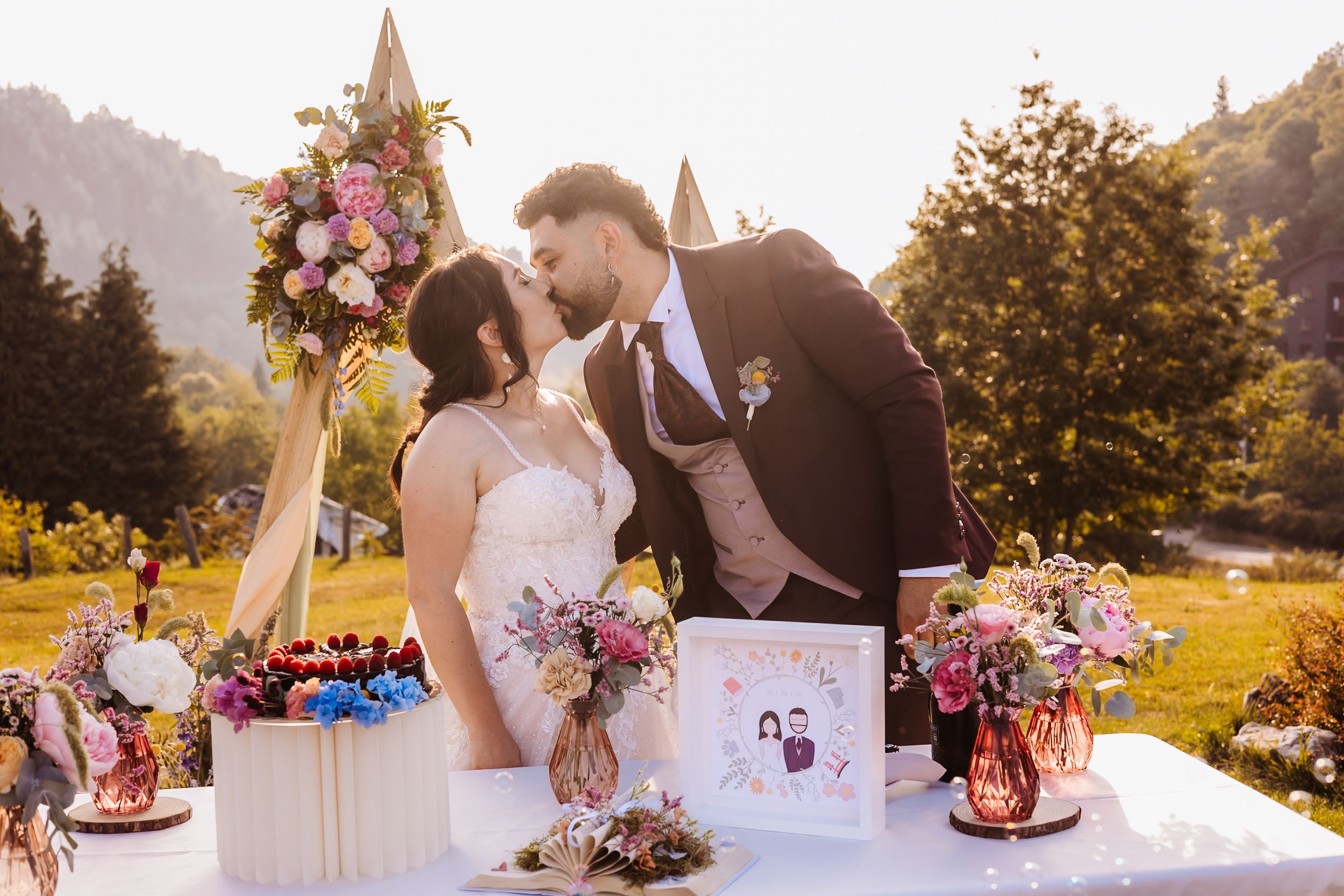 Bride and groom sharing a kiss at outdoor wedding ceremony with colorful floral arrangements and mountain backdrop.