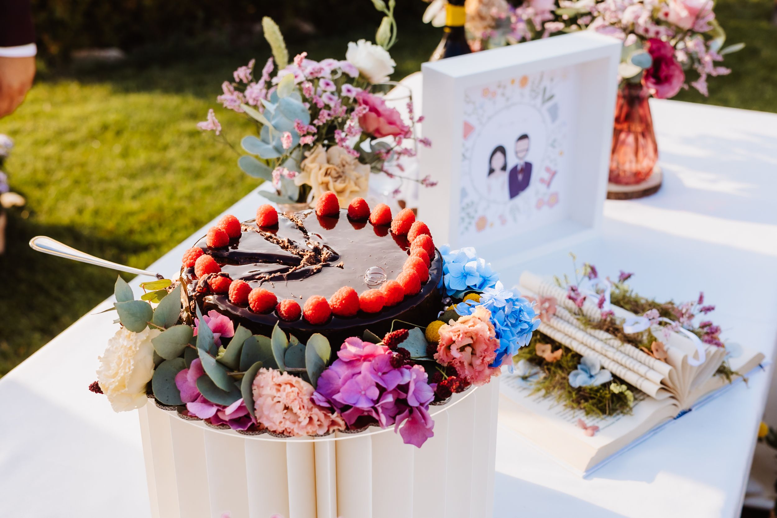 Elegant wedding cake topped with strawberries and chocolate, surrounded by colorful flowers on a white table outdoors.